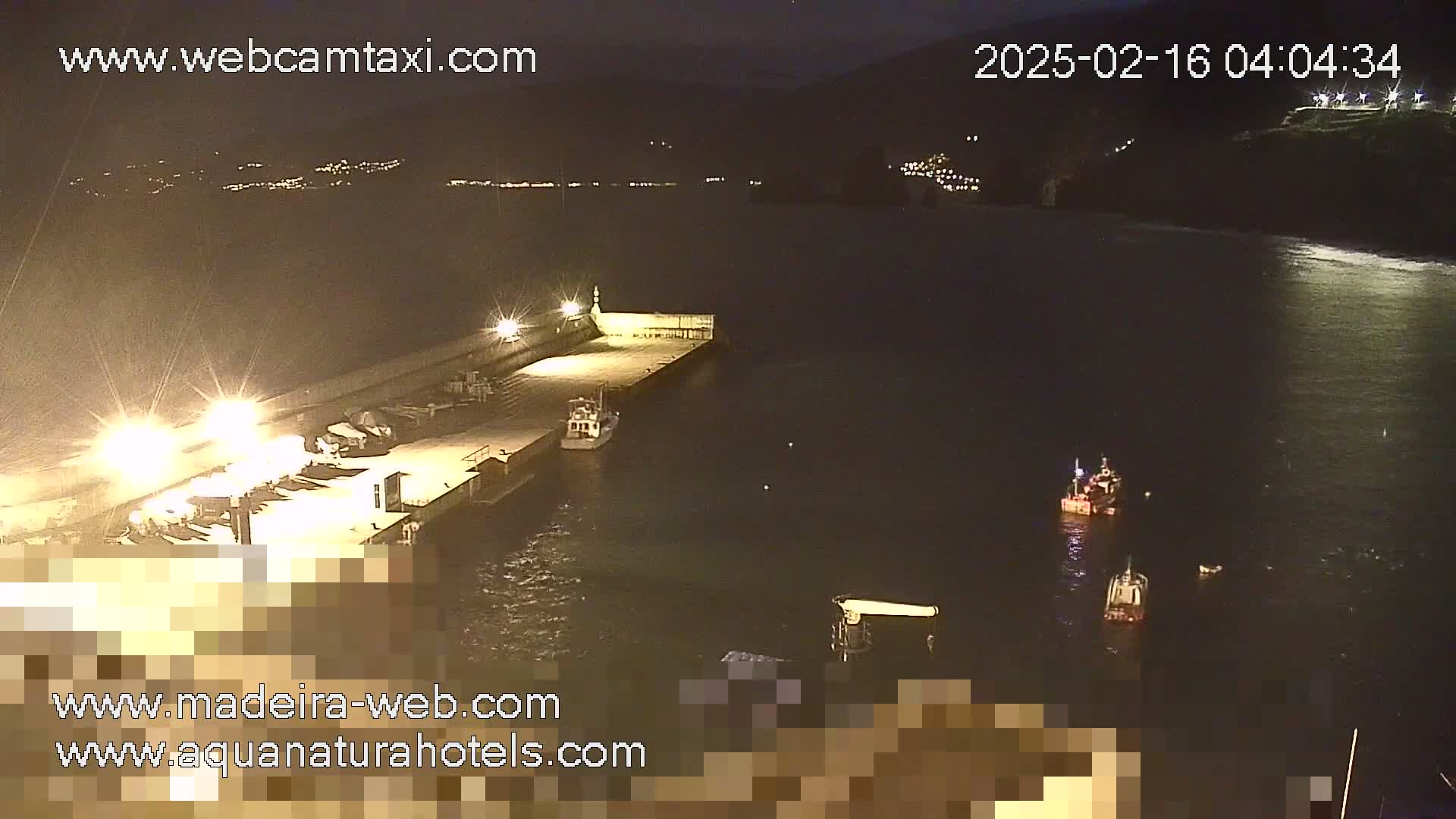 A nighttime view of a harbor with several small boats, one docked and others in the water, under a dark sky.