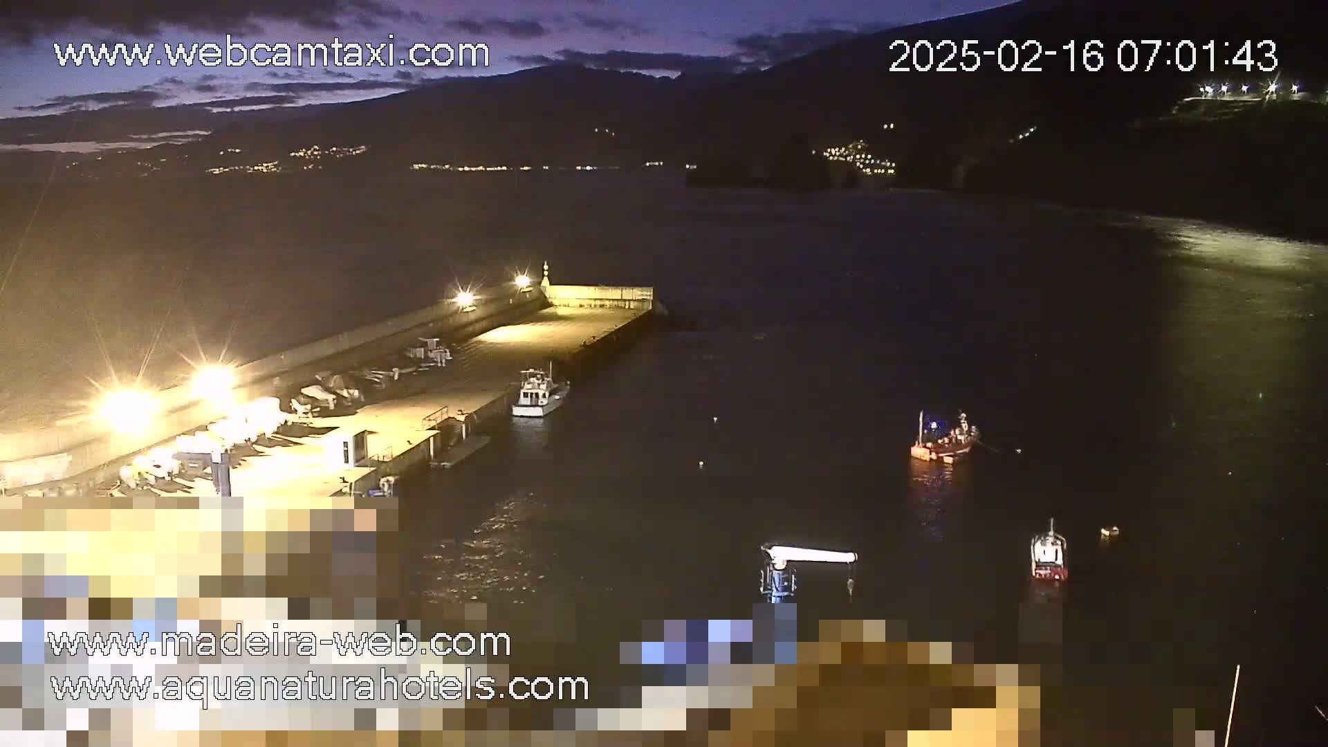 A nighttime harbor scene shows a pier with several boats moored, with additional boats in the dark water under a mostly clear sky.