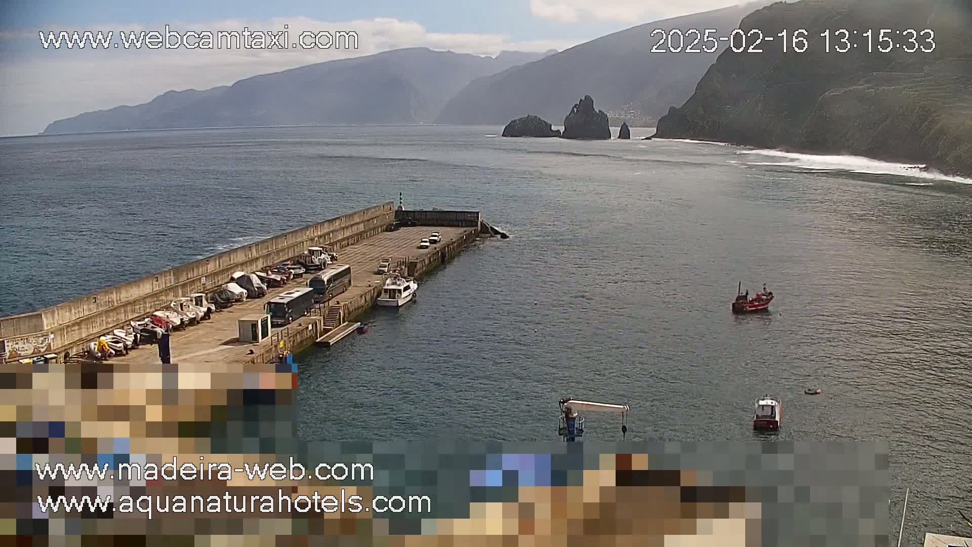 A small harbor with parked cars and boats is situated near a rocky coastline under partly sunny skies.
