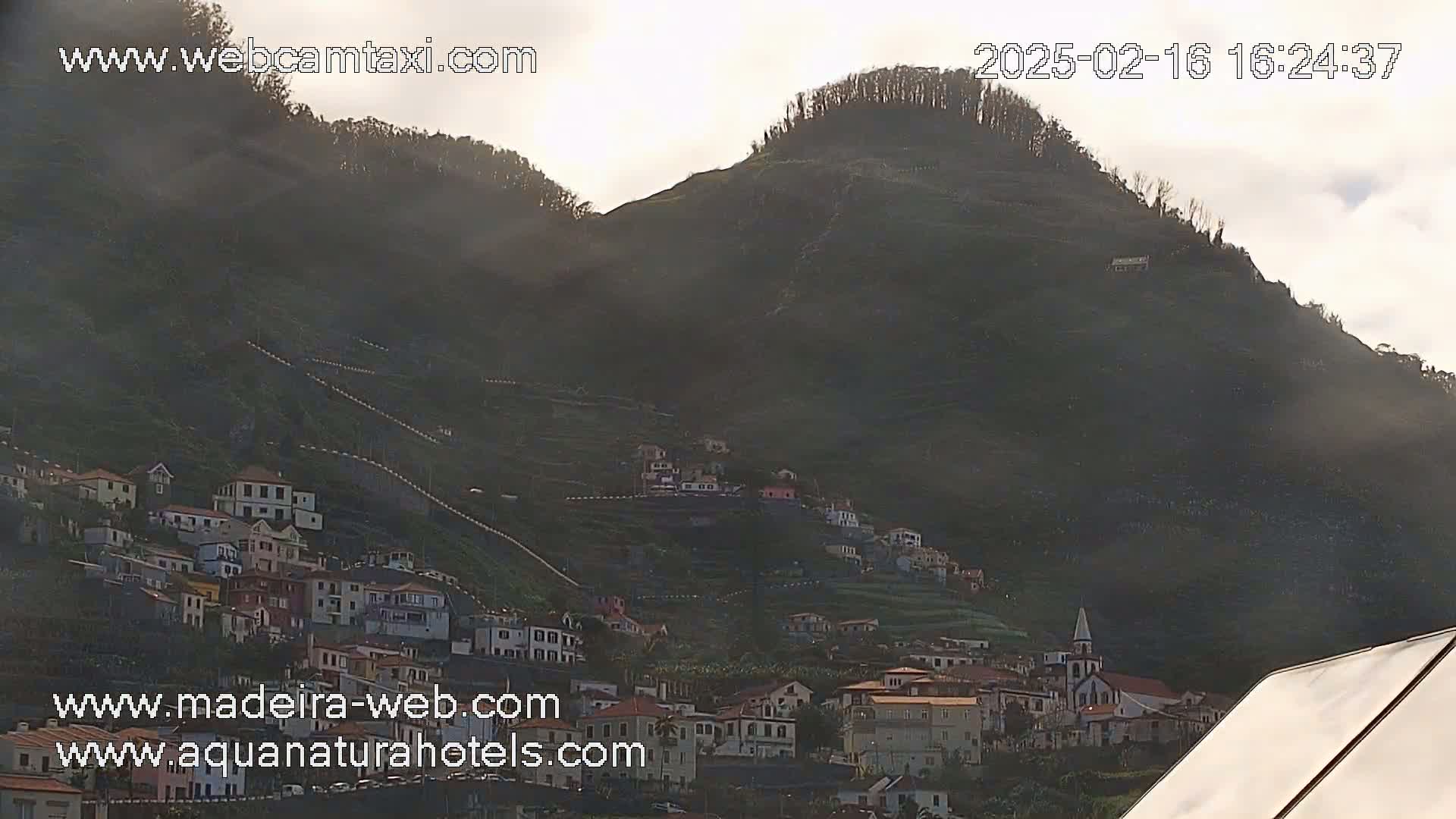 A hillside village clings to the slopes of a dark green mountain under a hazy, pale sky.
