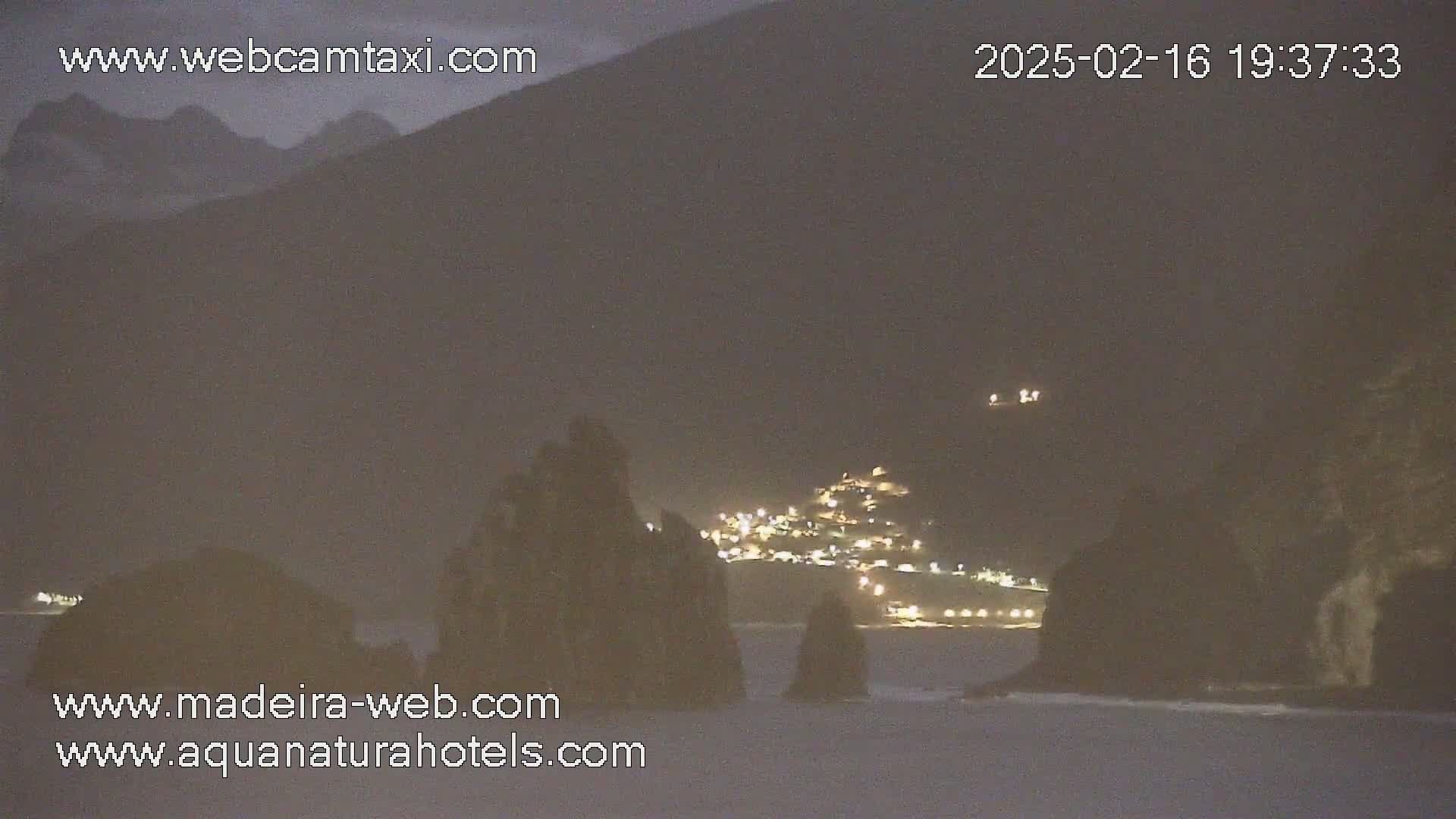A nighttime coastal scene shows dark, rocky outcroppings in the foreground with a faintly lit town nestled in the hills beyond under a hazy sky.