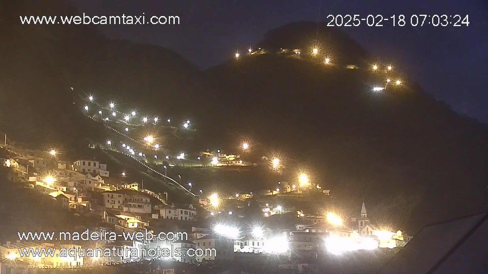 A nighttime view of a hillside village illuminated by numerous lights, climbing a dark mountain under a clear night sky.