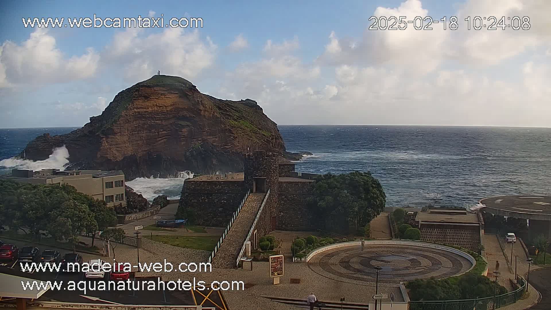 A stone structure with a walkway overlooks a rocky coast where powerful waves crash against the shore under a partly cloudy sky.