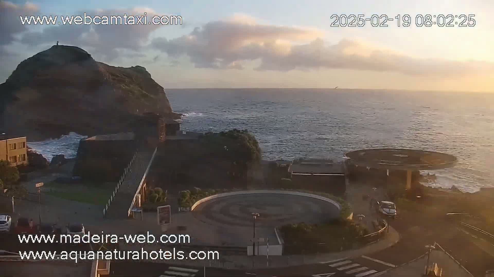 A coastal scene at sunrise shows a rocky outcrop with buildings nearby, a circular plaza, and calm ocean waves.