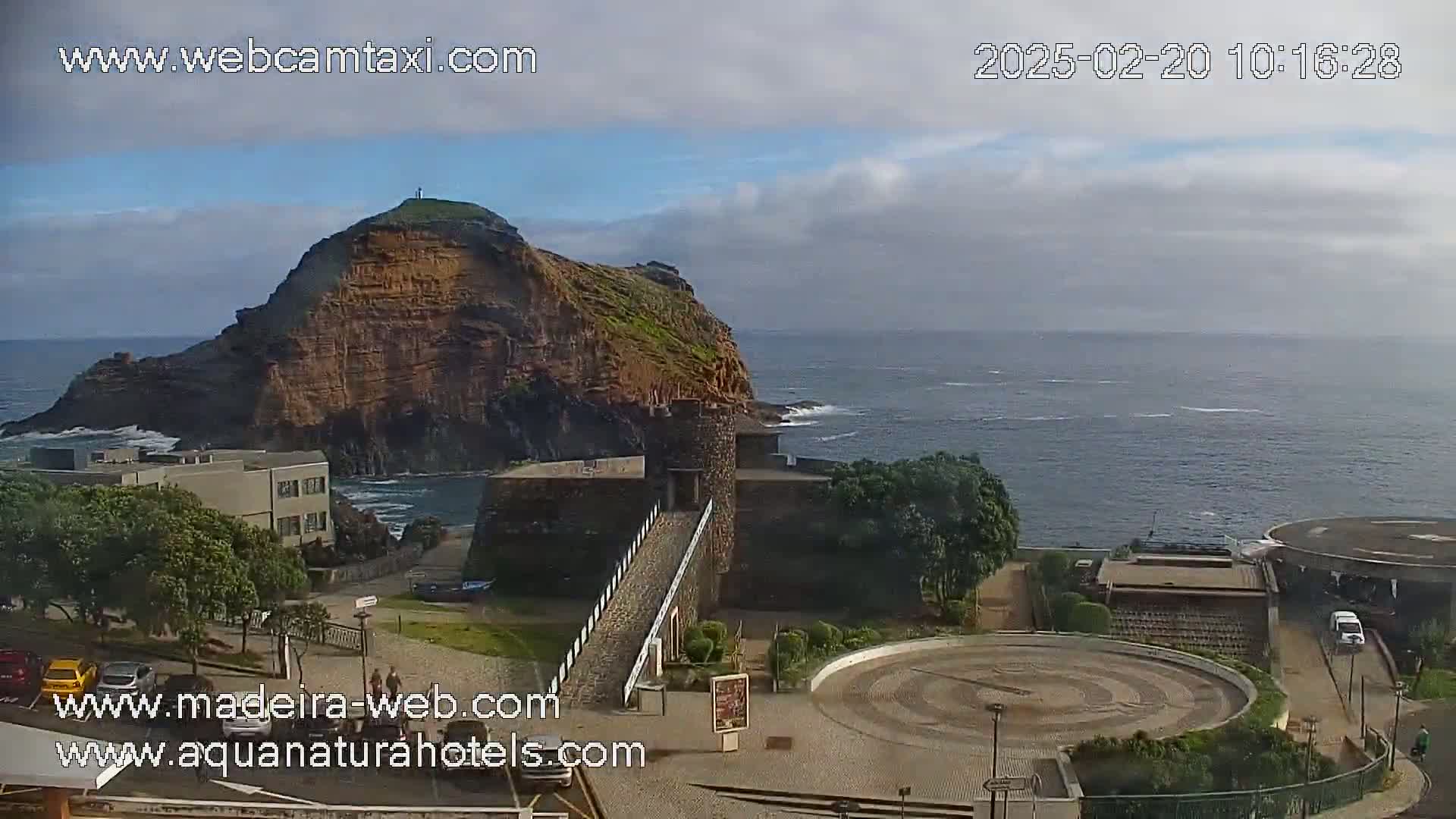 A coastal scene on a partly cloudy day shows a rocky hill, a stone structure with a walkway, and a circular plaza near the ocean.