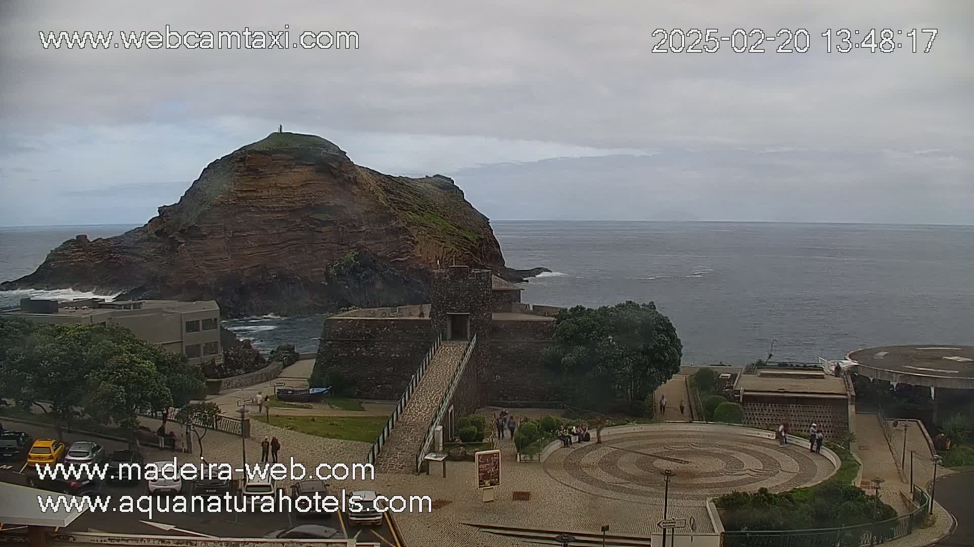 A rocky, reddish-brown island is visible in the ocean under an overcast sky, with a stone structure and surrounding buildings in the foreground.
