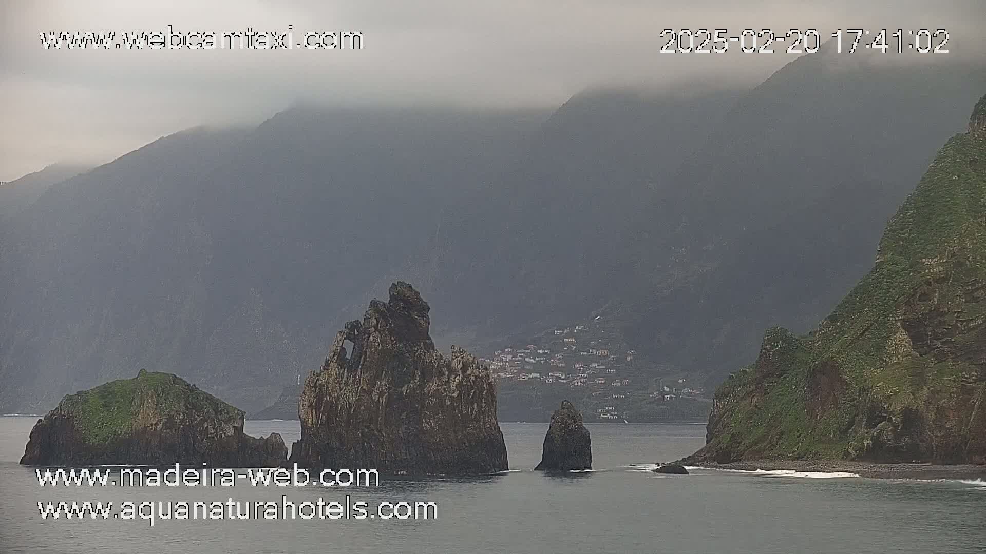 A hazy view shows several dark rock formations in the ocean near a mountainous coastline under an overcast sky.
