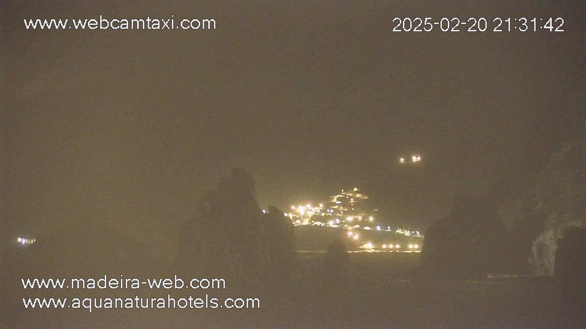 A nighttime long shot shows a dimly lit village nestled on a hillside, partially obscured by darkness and what appears to be fog or haze.