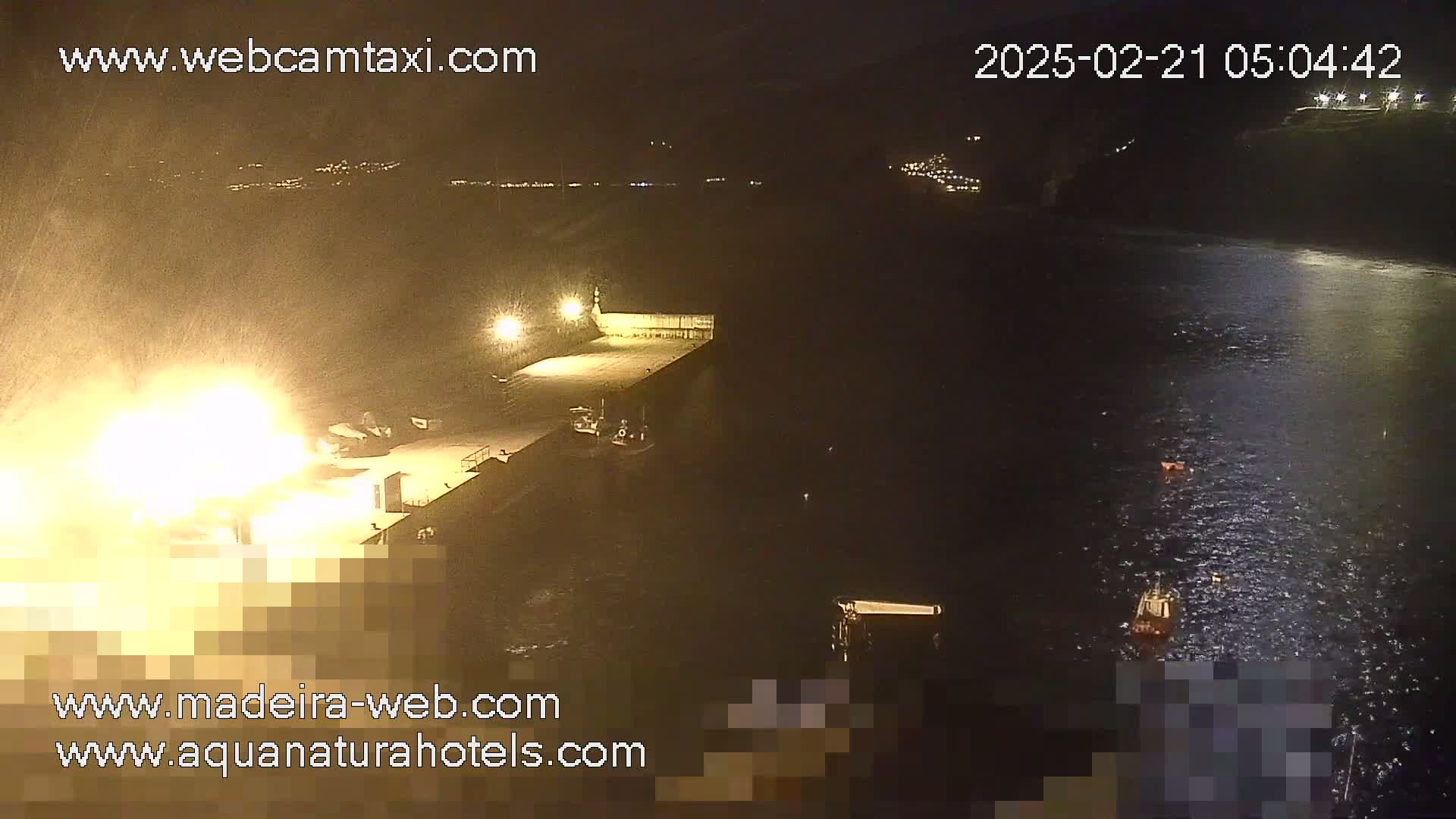 A nighttime, rain-swept view of a harbor shows a brightly lit dock with several boats and a large, bright light source possibly indicating a fire or explosion near the dock.