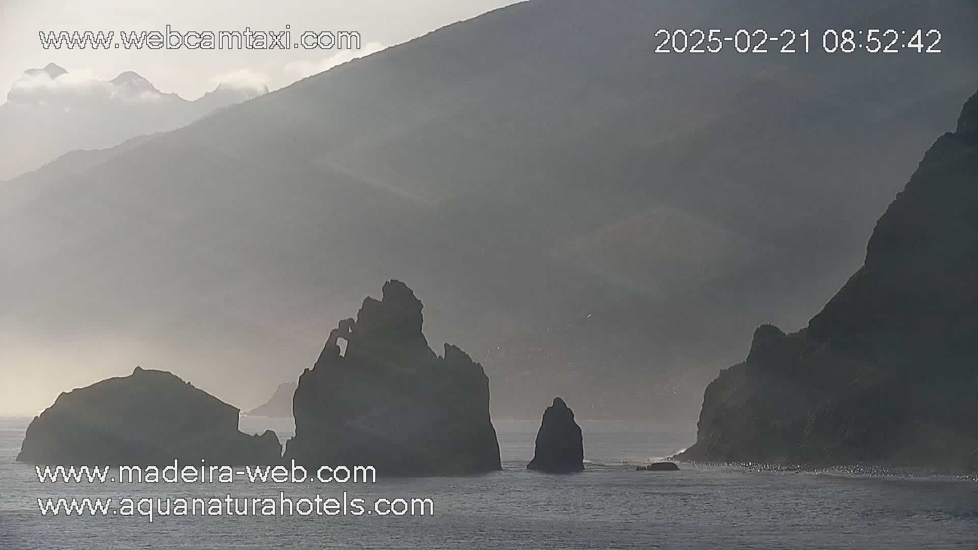 Silhouetted rocky outcrops stand in a calm sea near a hazy, mountainous coastline under a misty sky.
