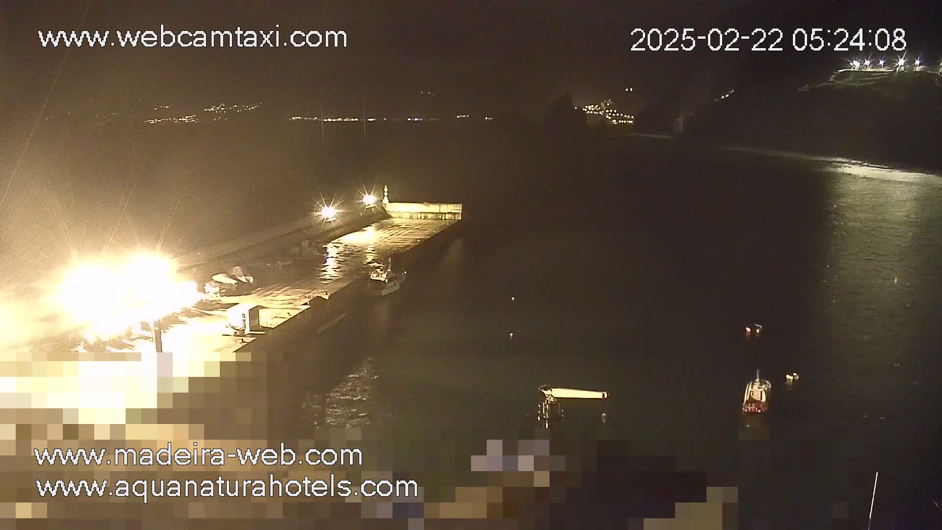 A nighttime, low-angle view shows a brightly lit dock with several boats moored alongside, and a few other boats are visible in dark water beyond.