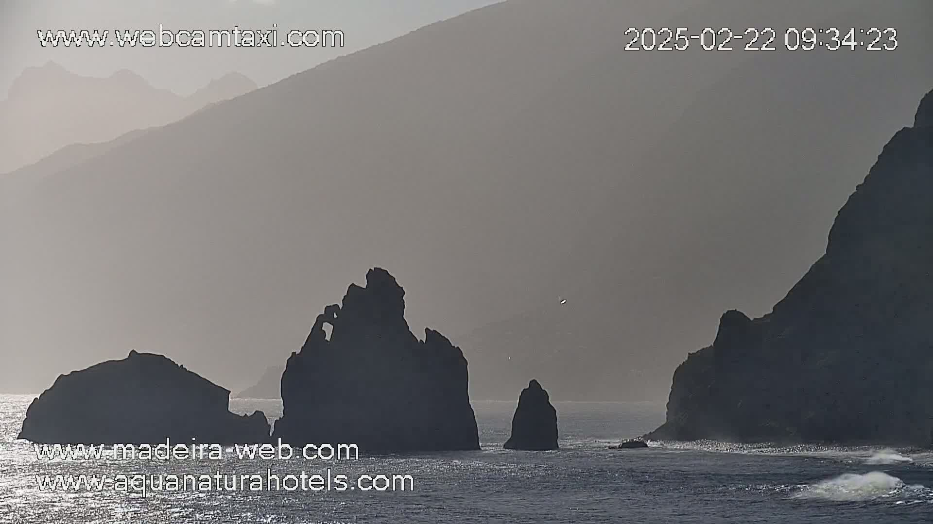 Silhouetted rocky outcrops and distant mountains are seen in hazy, overcast conditions over a dark body of water.