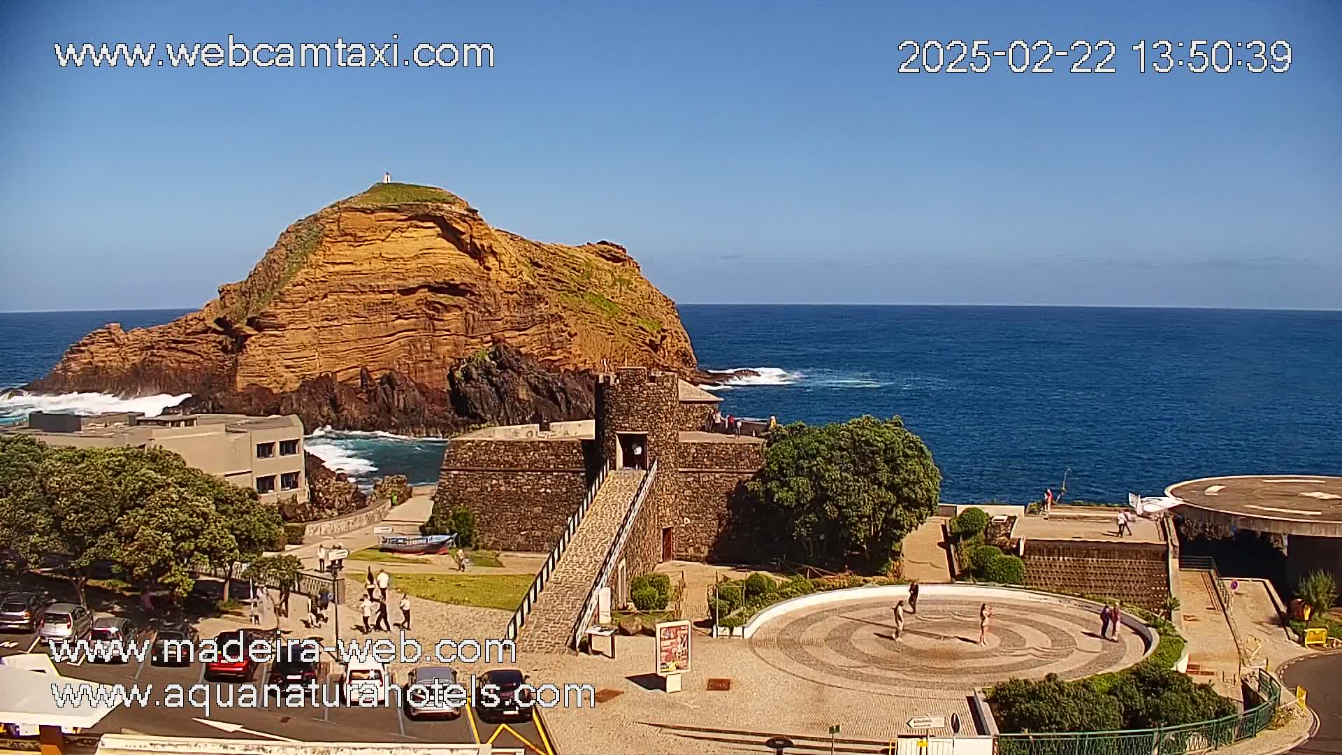 A rocky, orange-brown island sits in a blue ocean under a clear, sunny sky, with a stone structure and a paved plaza visible on the shore.