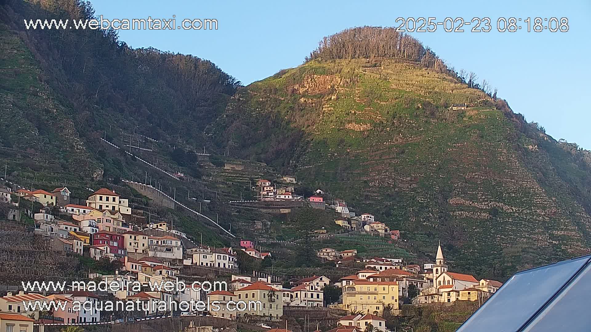 A hillside village clings to the slopes of a verdant mountain under a clear, bright sky.