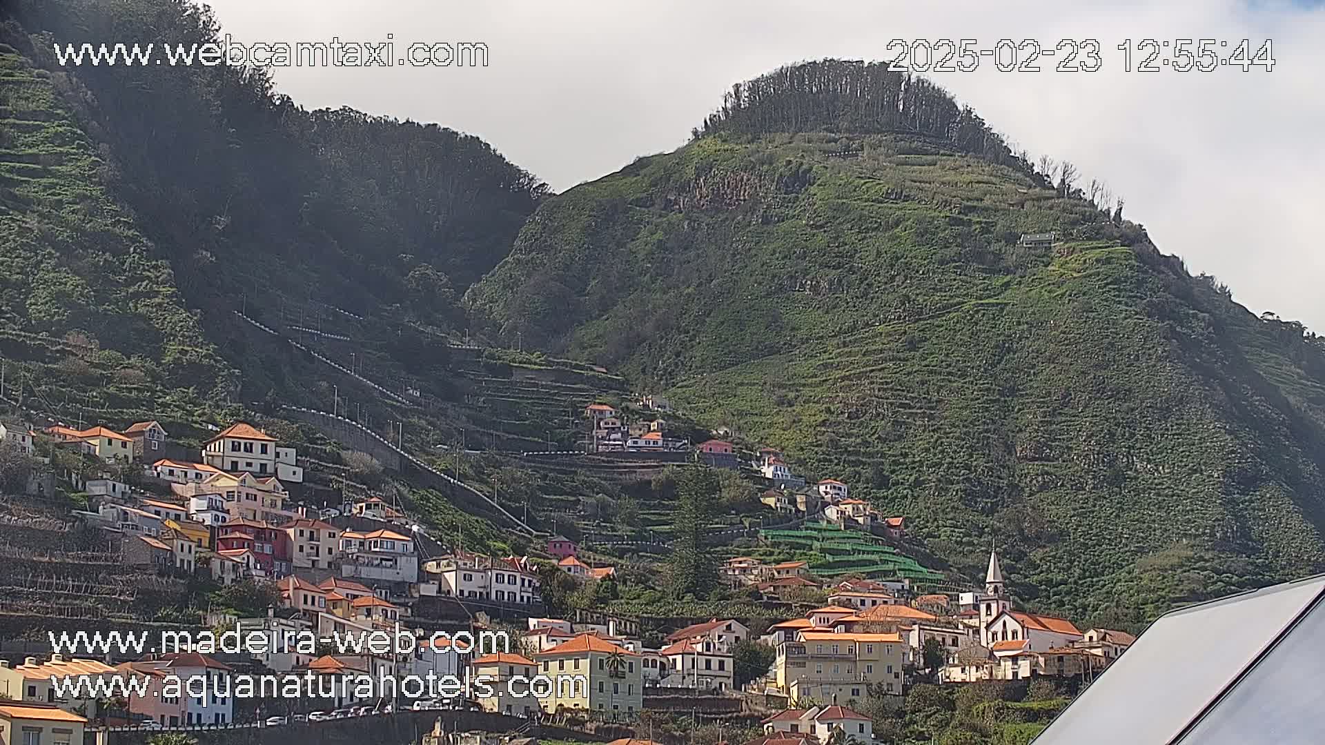 A village of red-roofed houses clings to the slopes of a verdant, terraced hillside under a mostly clear sky.