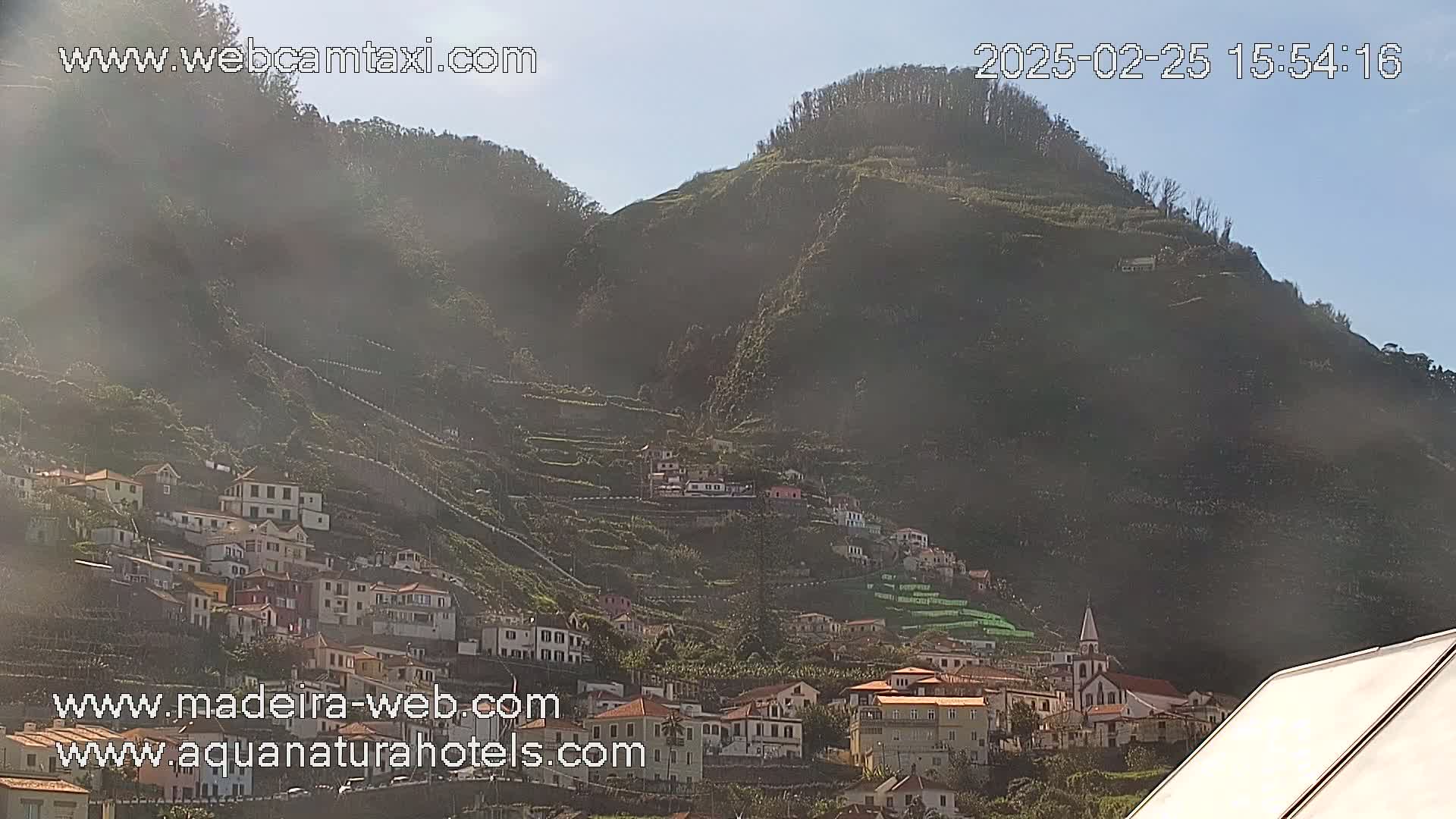 A village clings to the slopes of a hazy, sunlit green mountain.