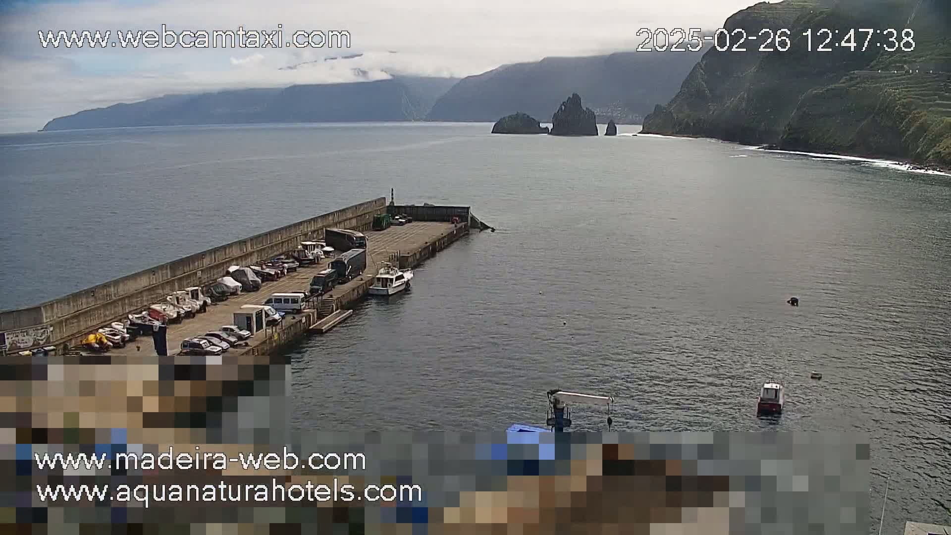 A small harbor with parked cars and boats is situated near a rocky coastline on a partly cloudy day.