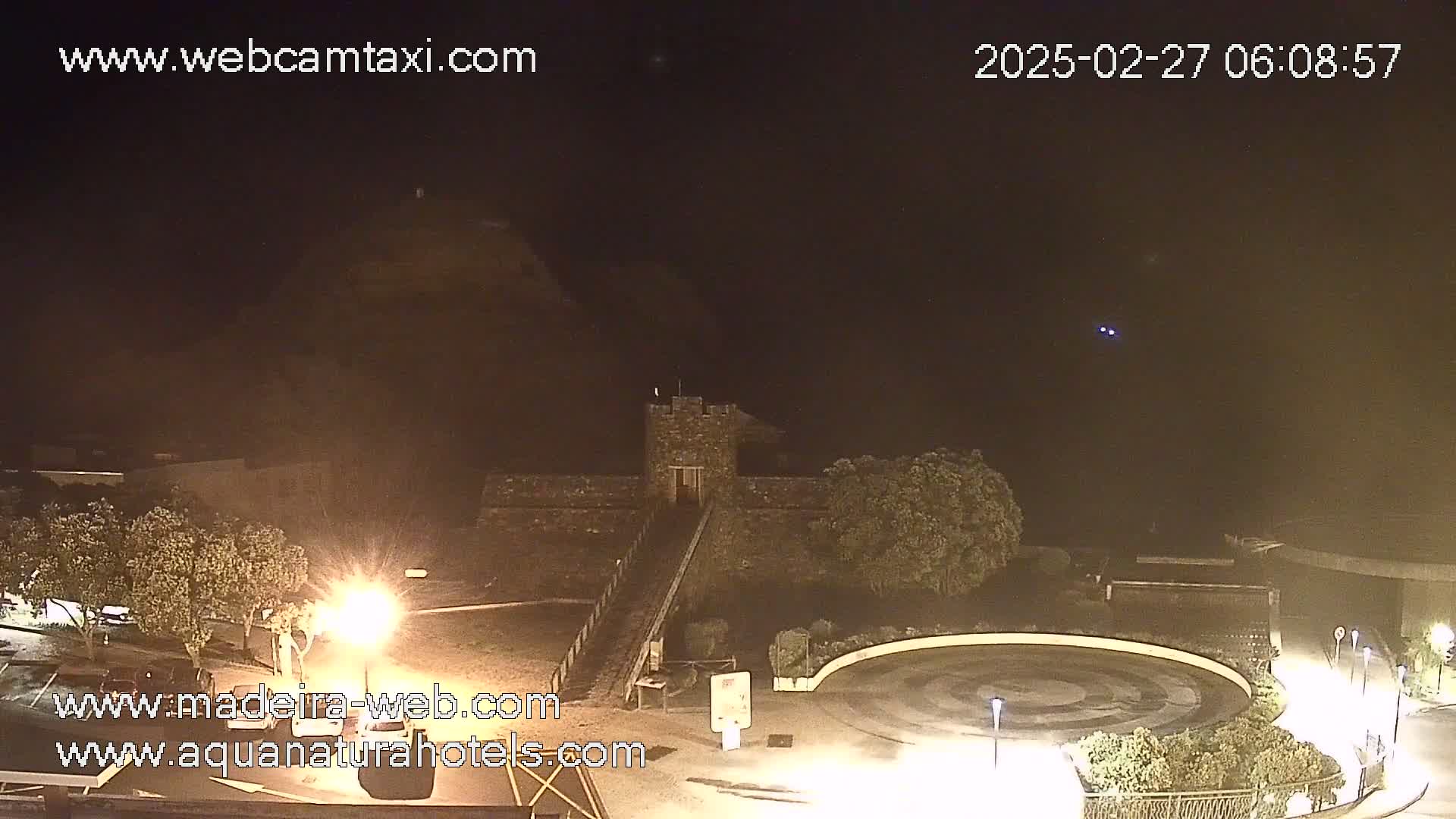 A nighttime, dimly lit view of a stone structure resembling a small castle, situated near a circular paved area with landscaping and some parked cars visible in the distance under a hazy, dark sky.