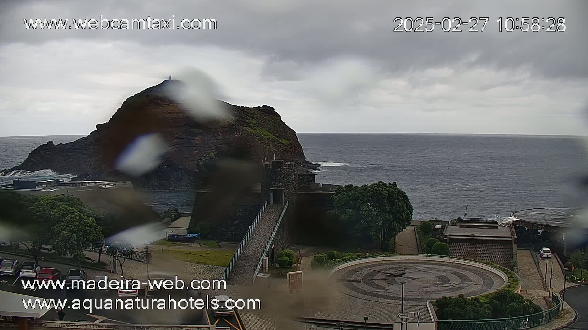 A rain-covered view shows a rocky coastal headland with a small building near the ocean, a circular plaza, and a parking lot.