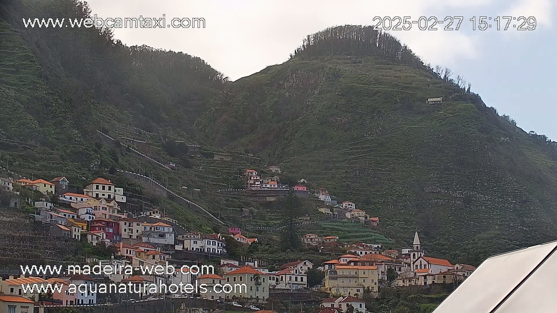 A hillside village with red-roofed houses climbs a steep, green mountain under an overcast sky.