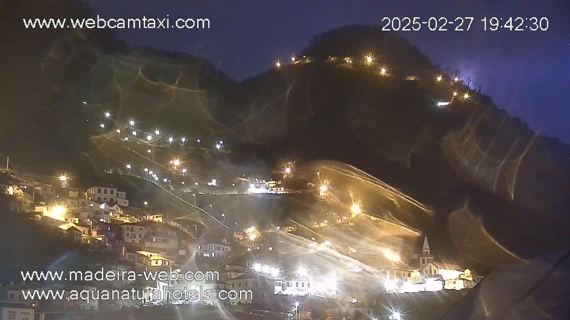 A nighttime view of a snow-covered hillside town illuminated by numerous lights, with some snow appearing to be blowing in the wind.