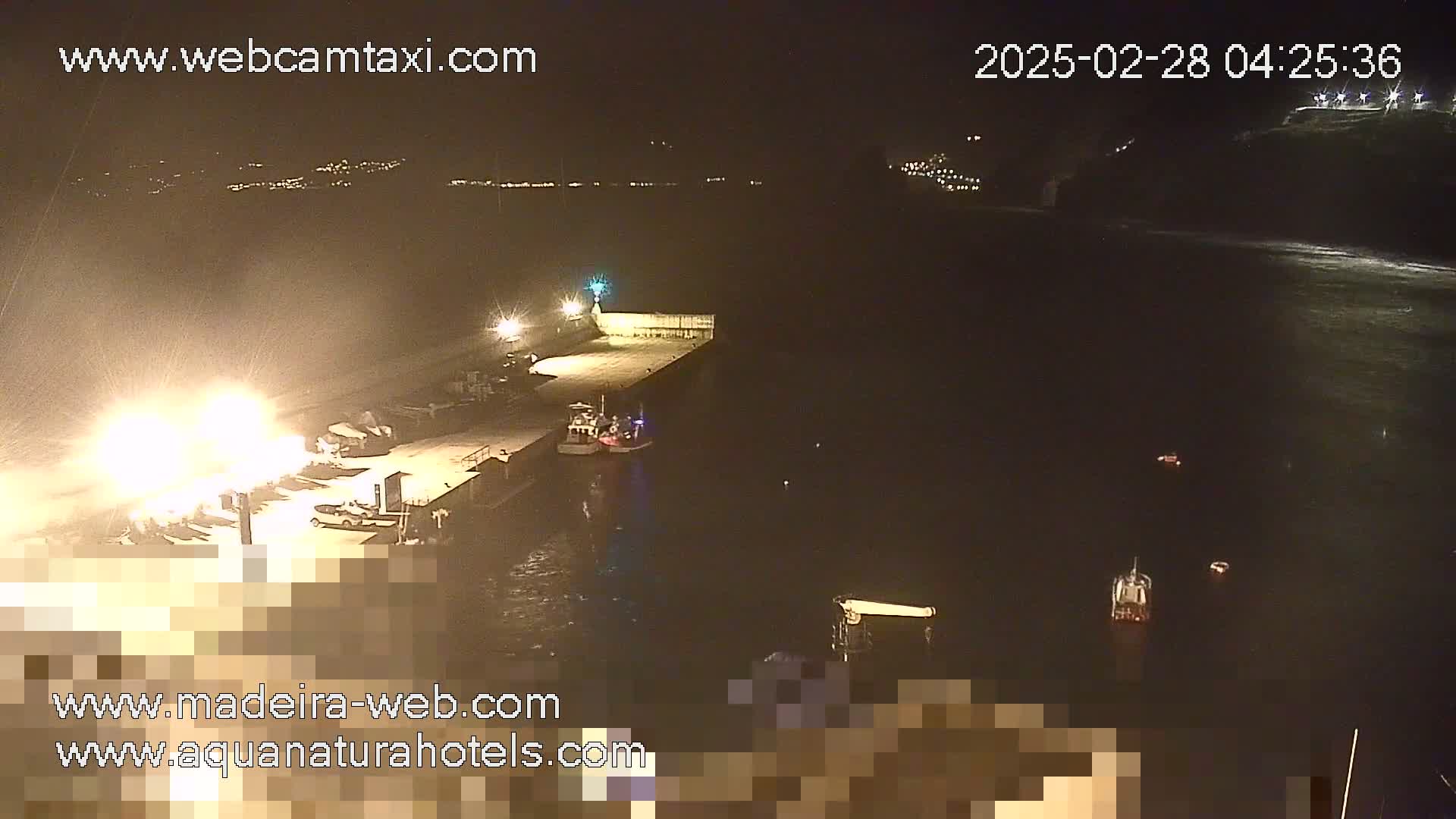 A nighttime, slightly overcast view of a harbor with several boats moored at a pier, and a few more vessels in the water.