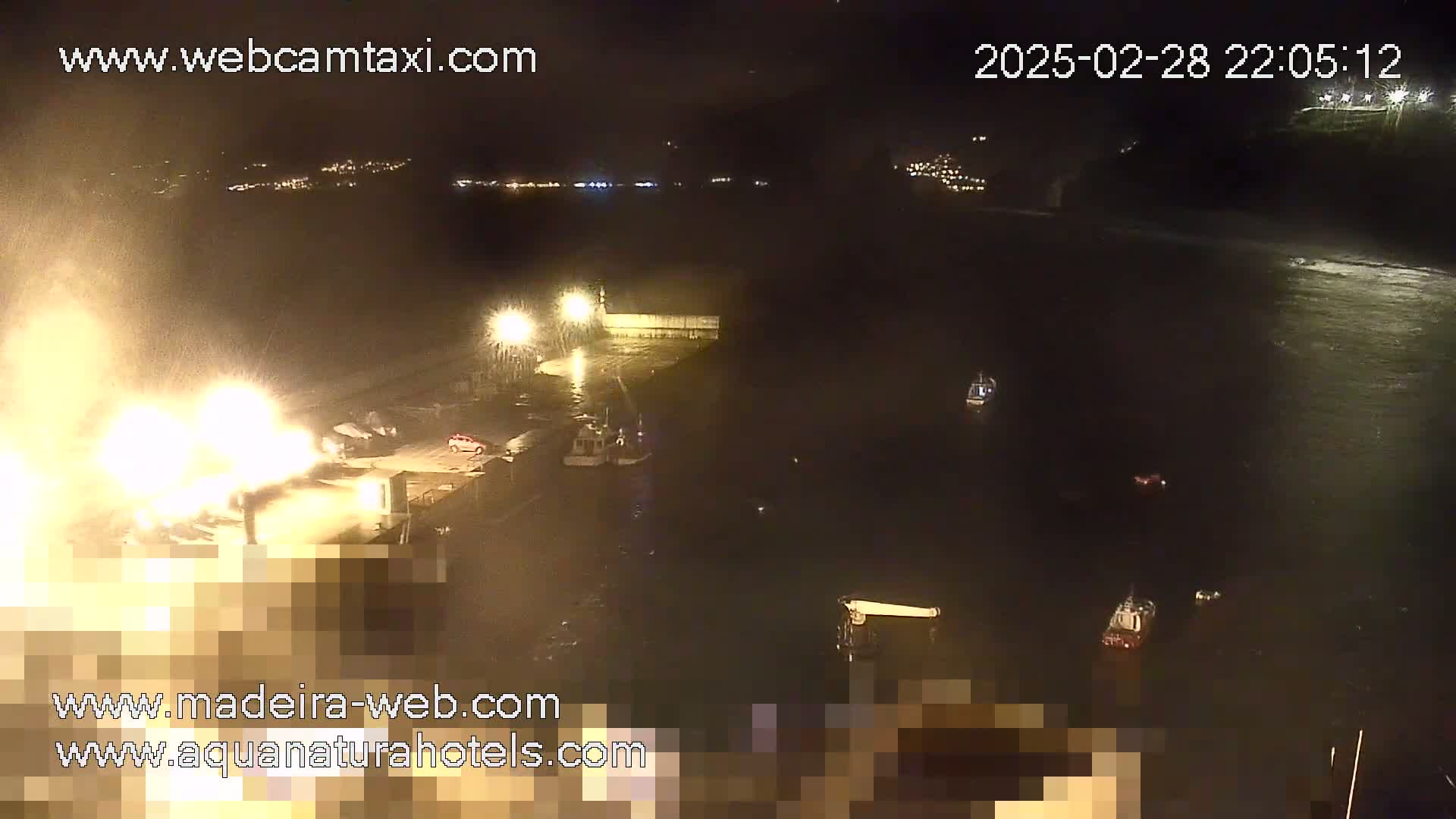A nighttime harbor scene with several boats, illuminated by lights on the dock, shows bright splashing water near the dock, possibly due to strong waves or a fire.