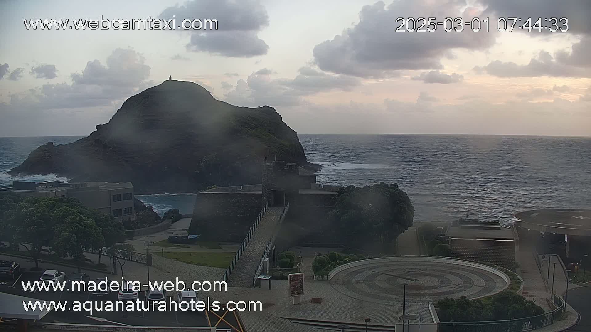 A coastal scene with overcast skies shows a dark rocky hill, a stone structure near the ocean, and a paved area with a circular design in the foreground, under partly cloudy skies.