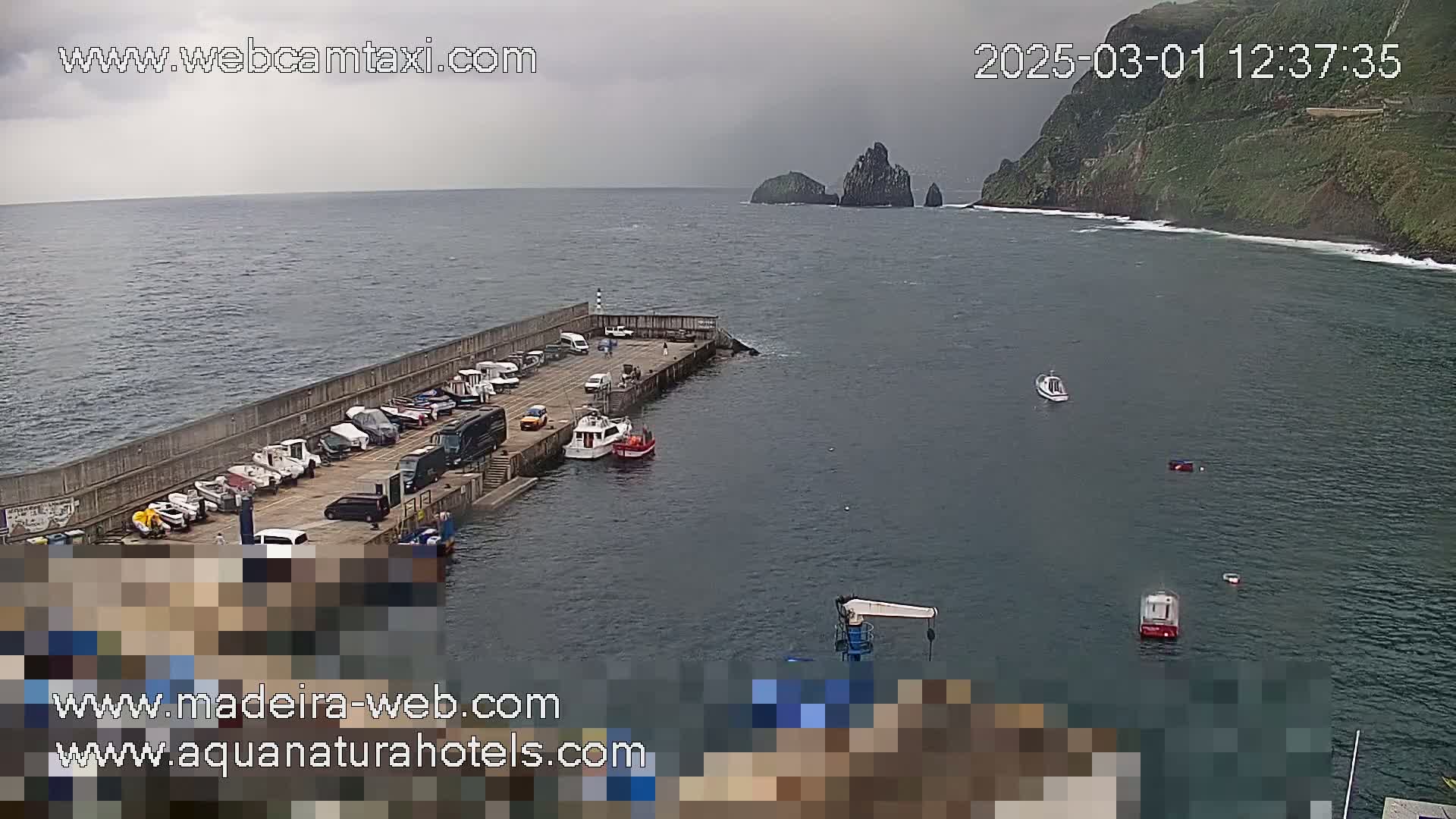 A small harbor with several parked cars and boats, situated on a calm, overcast day next to a steep, green cliffside.