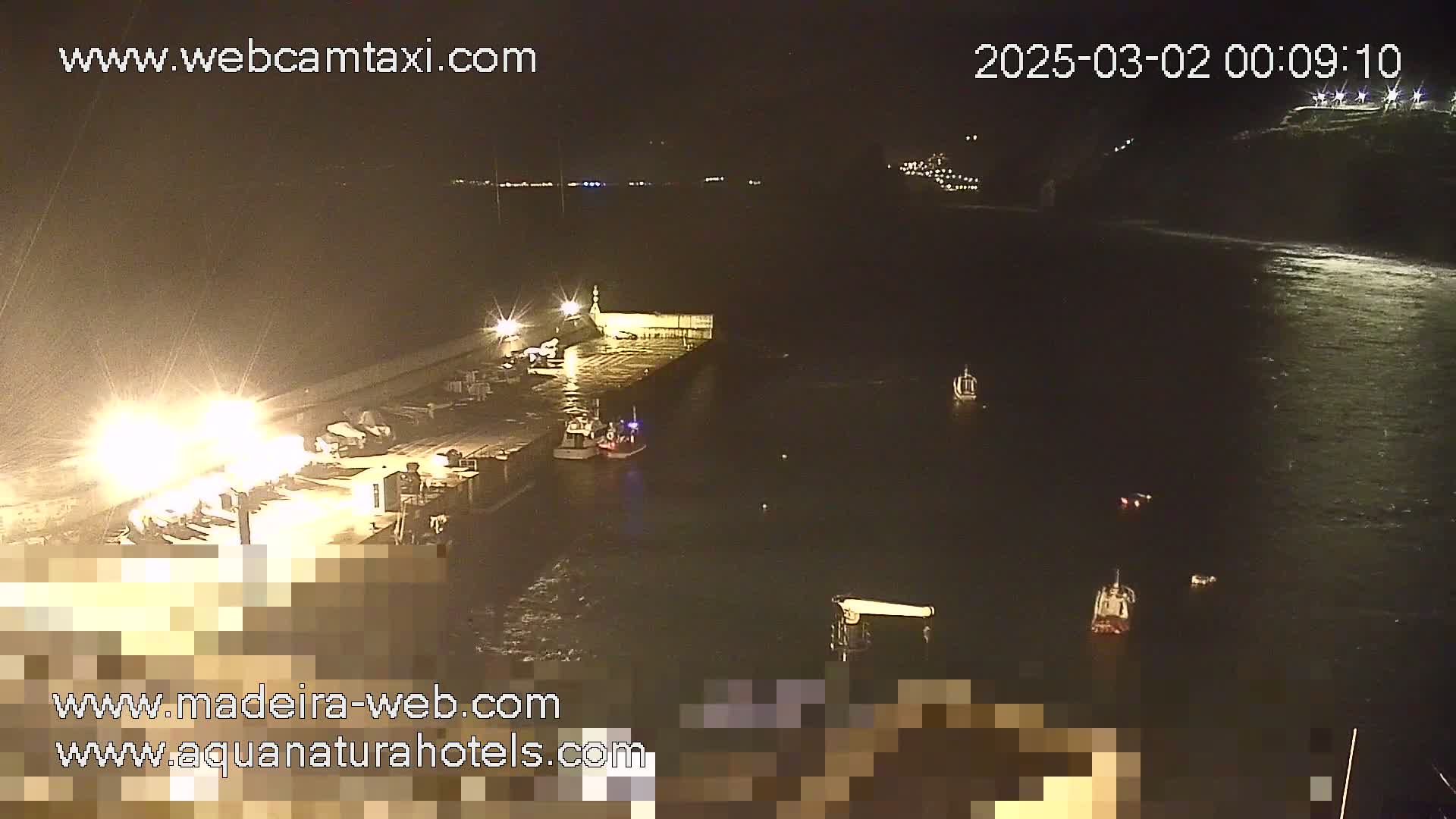 At night, several small boats are moored near a brightly lit dock next to a dark body of water under a clear sky.