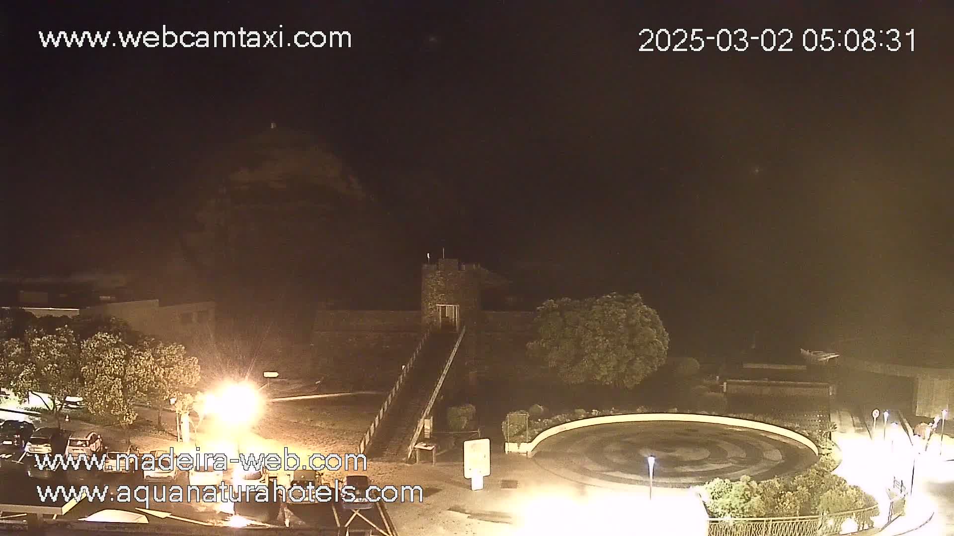 A nighttime, hazy view shows a stone structure with a ramp leading to it, surrounded by trees and a circular paved area with lights.