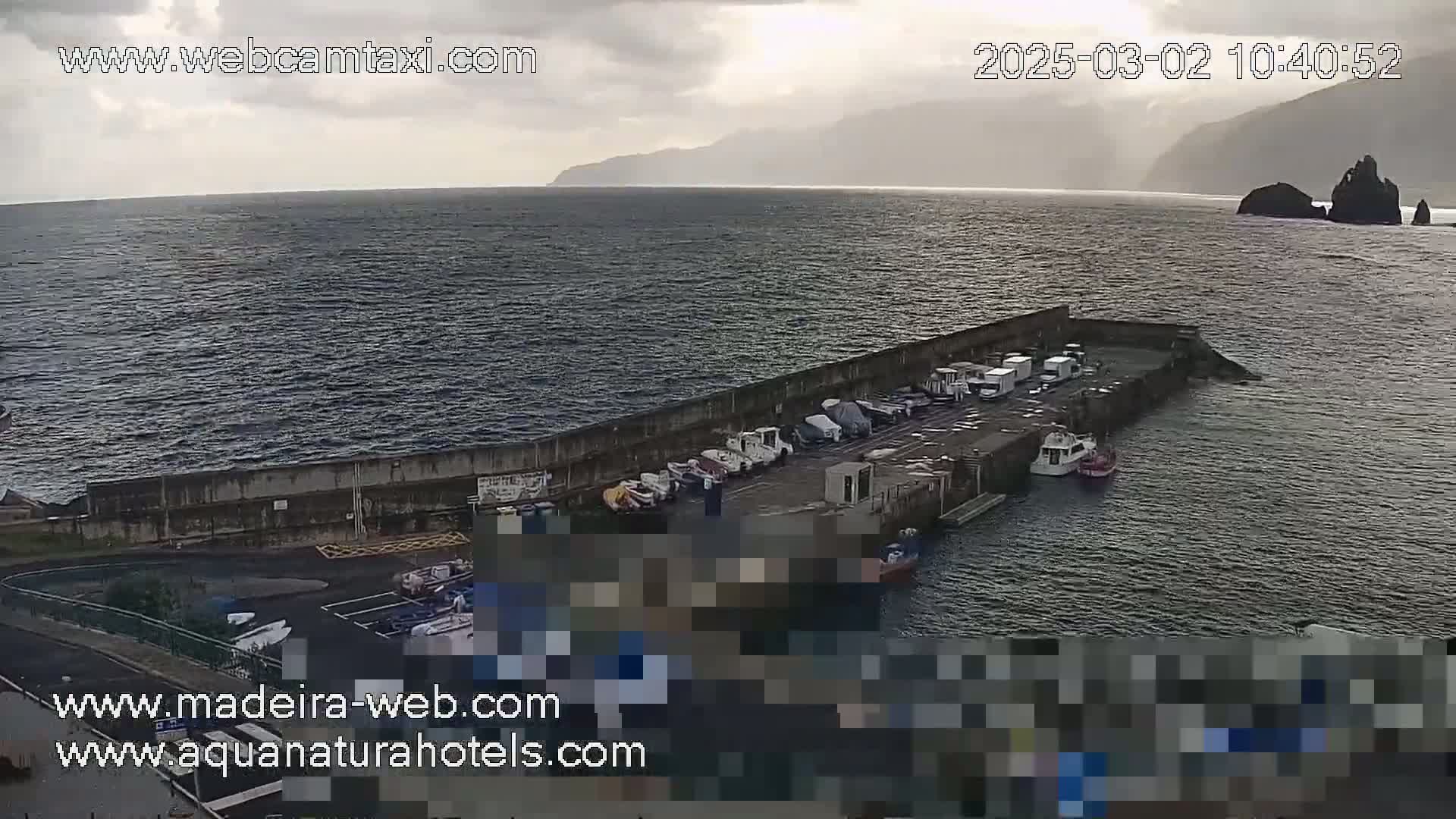 A small harbor with boats and vehicles parked on a pier, situated beside a dark, choppy ocean under an overcast sky.