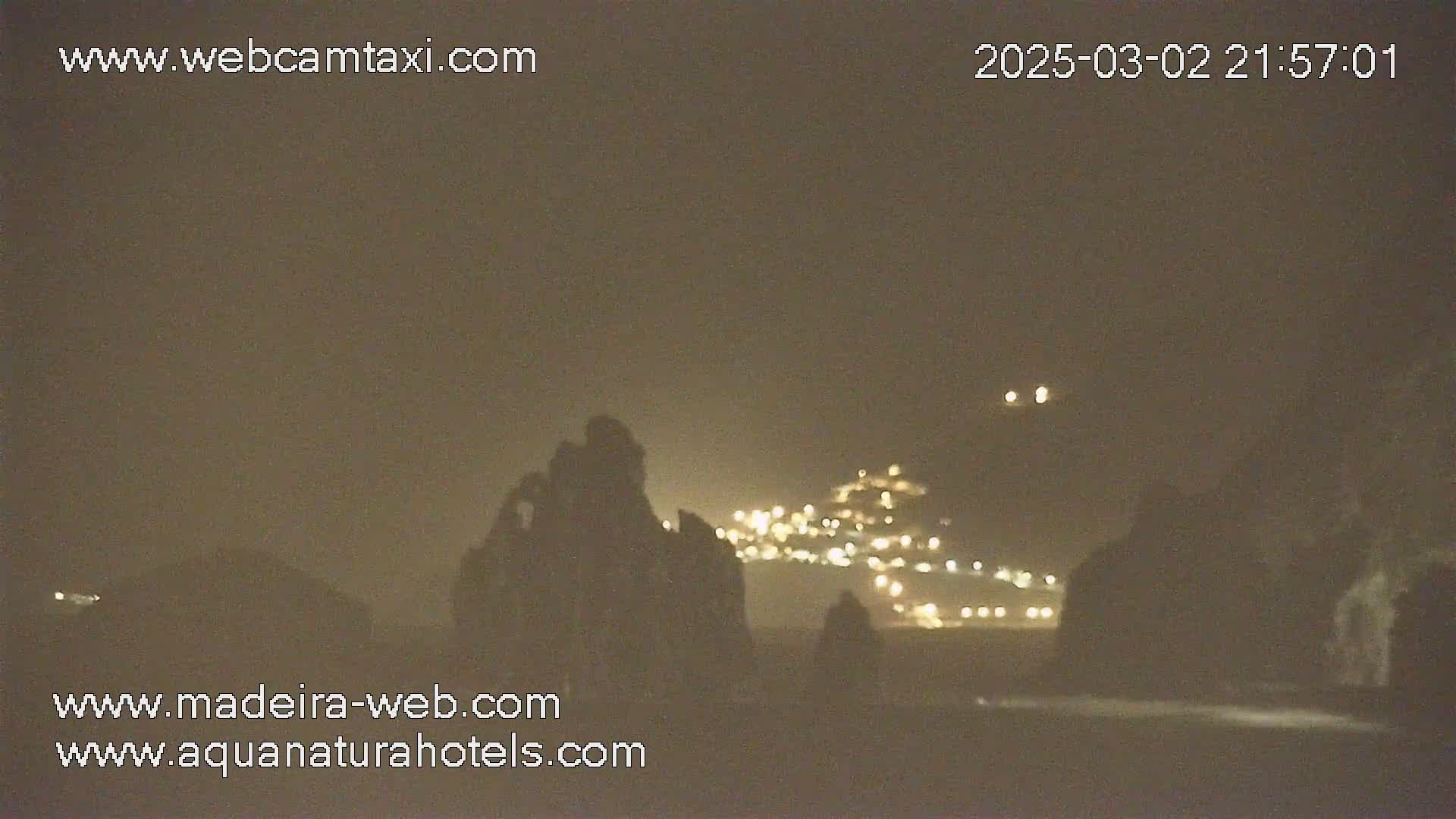 A nighttime, hazy view shows dark, silhouetted rock formations in the foreground with a blurry, lit town in the background.