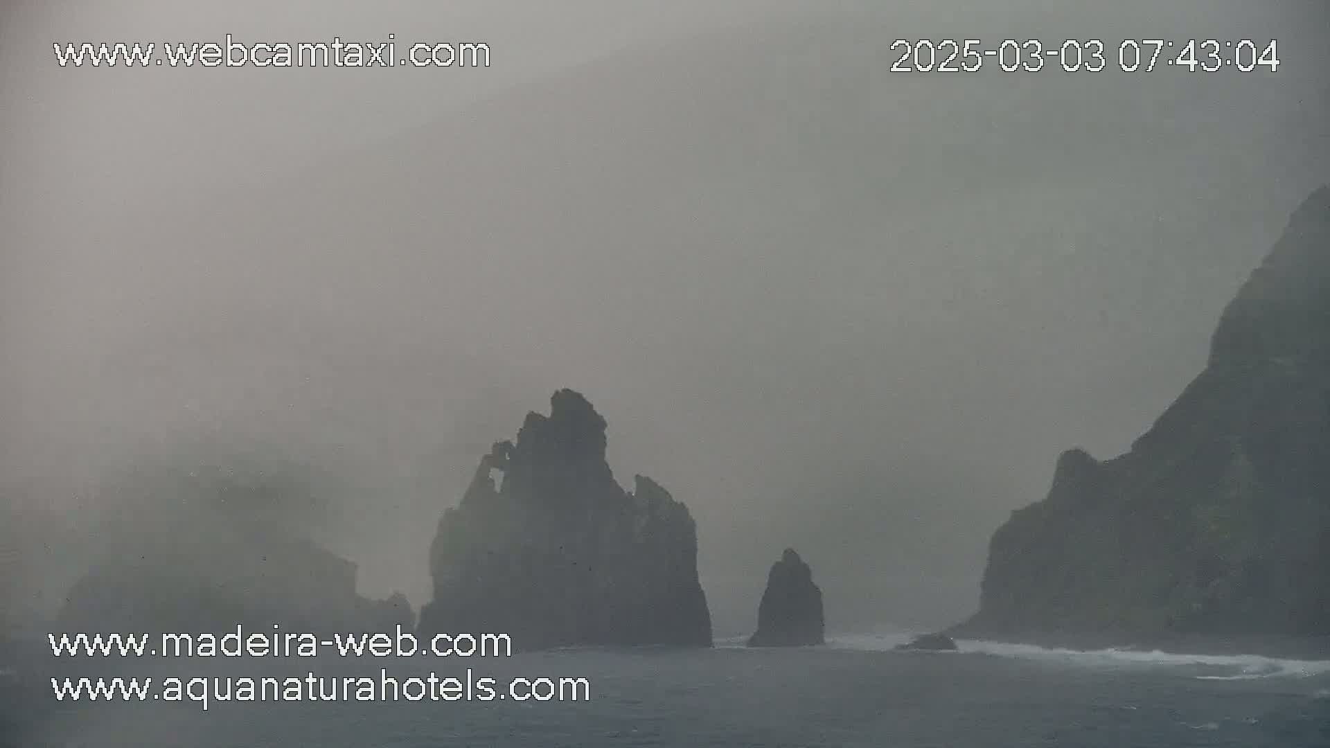 Dark, rocky sea stacks and cliffs are shrouded in thick fog over a choppy sea.