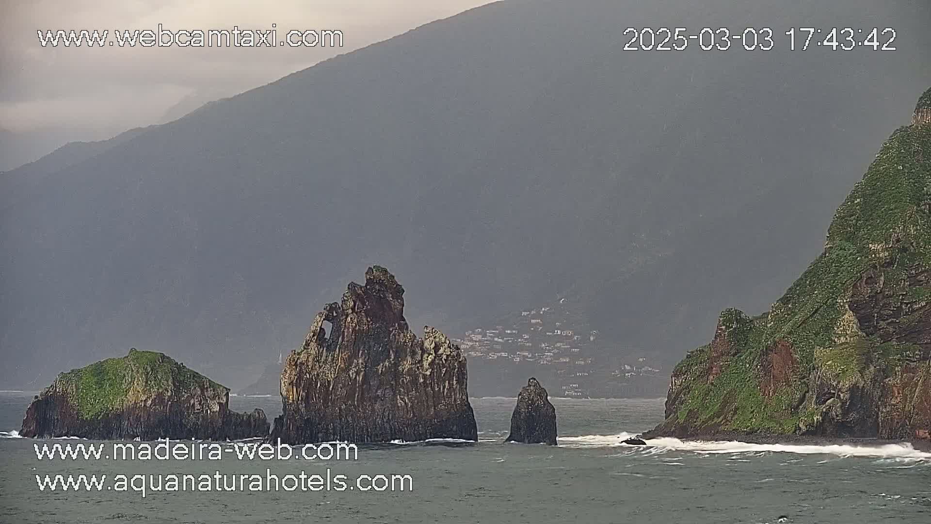 Several rocky outcroppings and small islands rise from a dark-grey sea under an overcast sky near a distant, hazy mountain range.