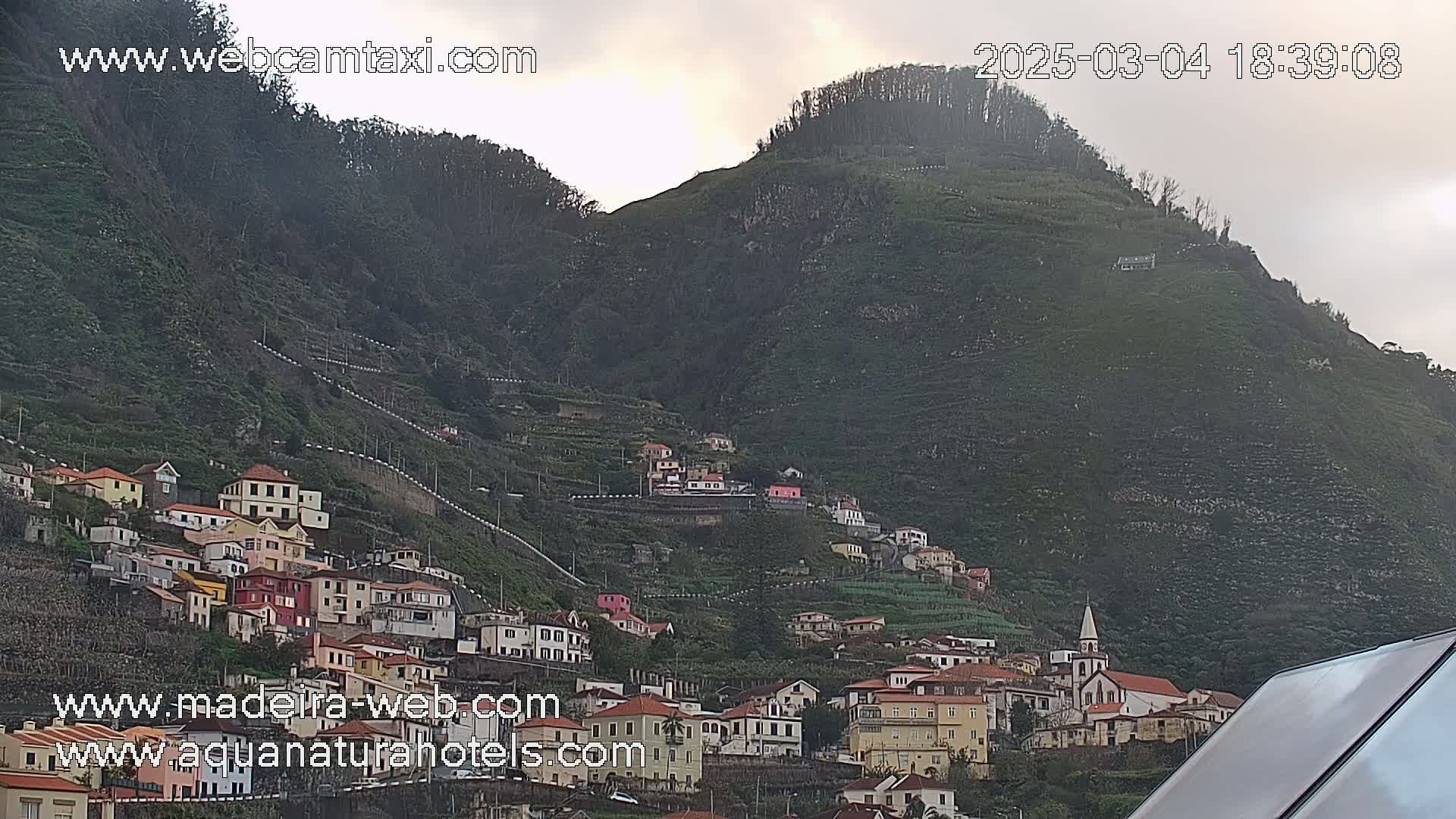 A hillside village of colorful buildings clings to a steep, verdant mountain under an overcast sky.
