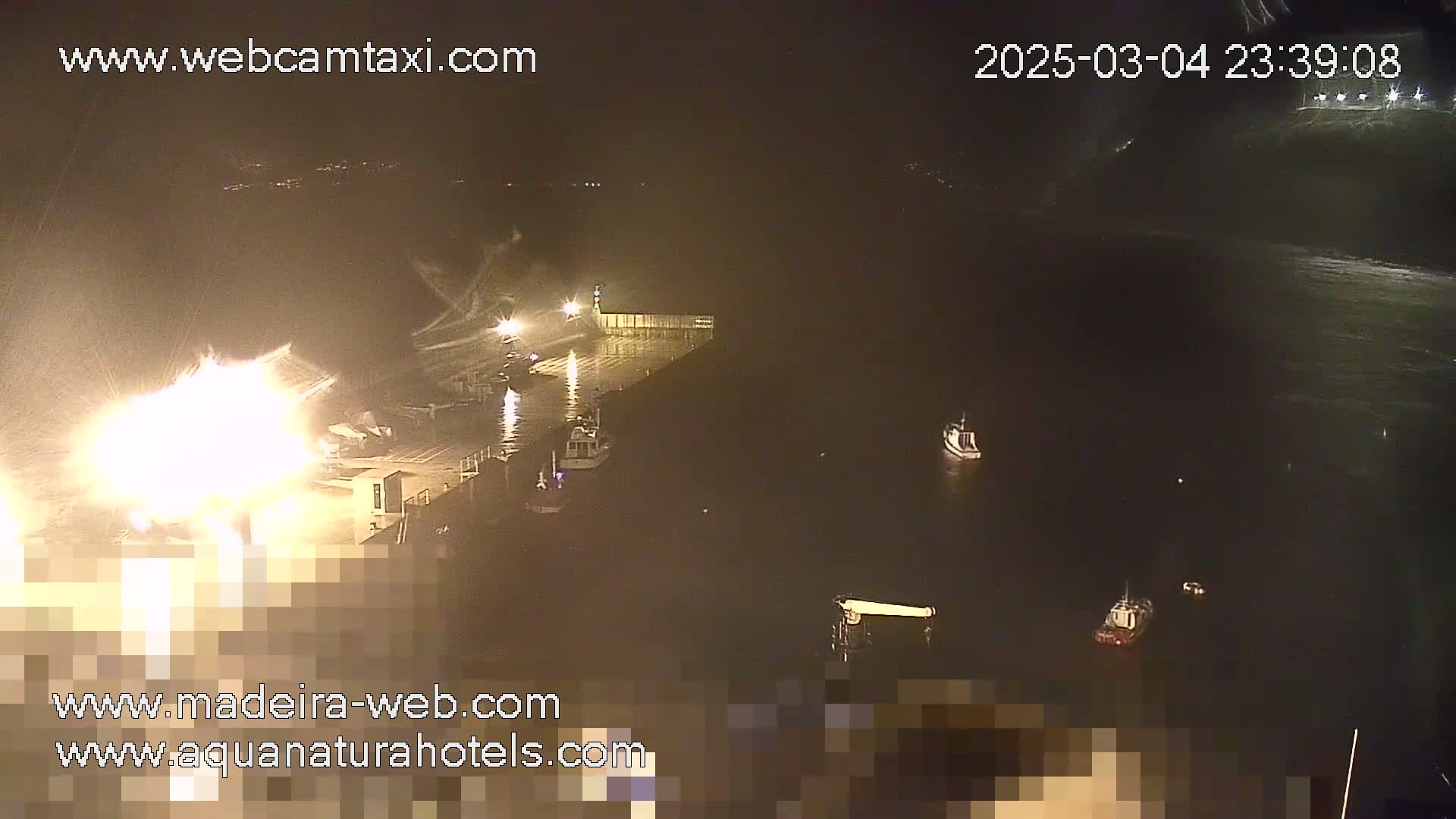 A nighttime, low-light view of a harbor with several boats moored, illuminated by bright lights on a pier.
