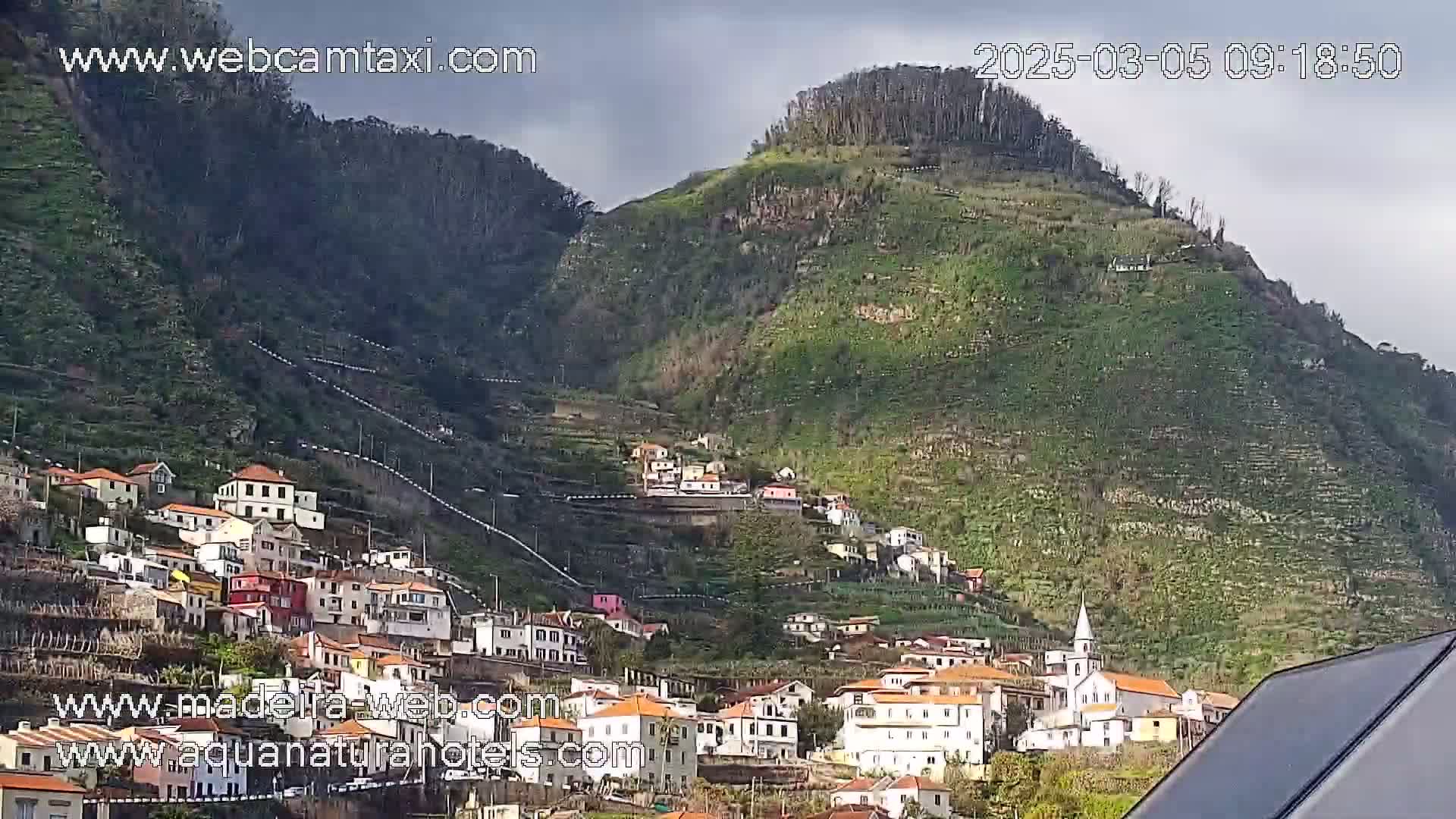A hillside village with terraced farmland and a church, nestled on a steep mountainside under an overcast sky.