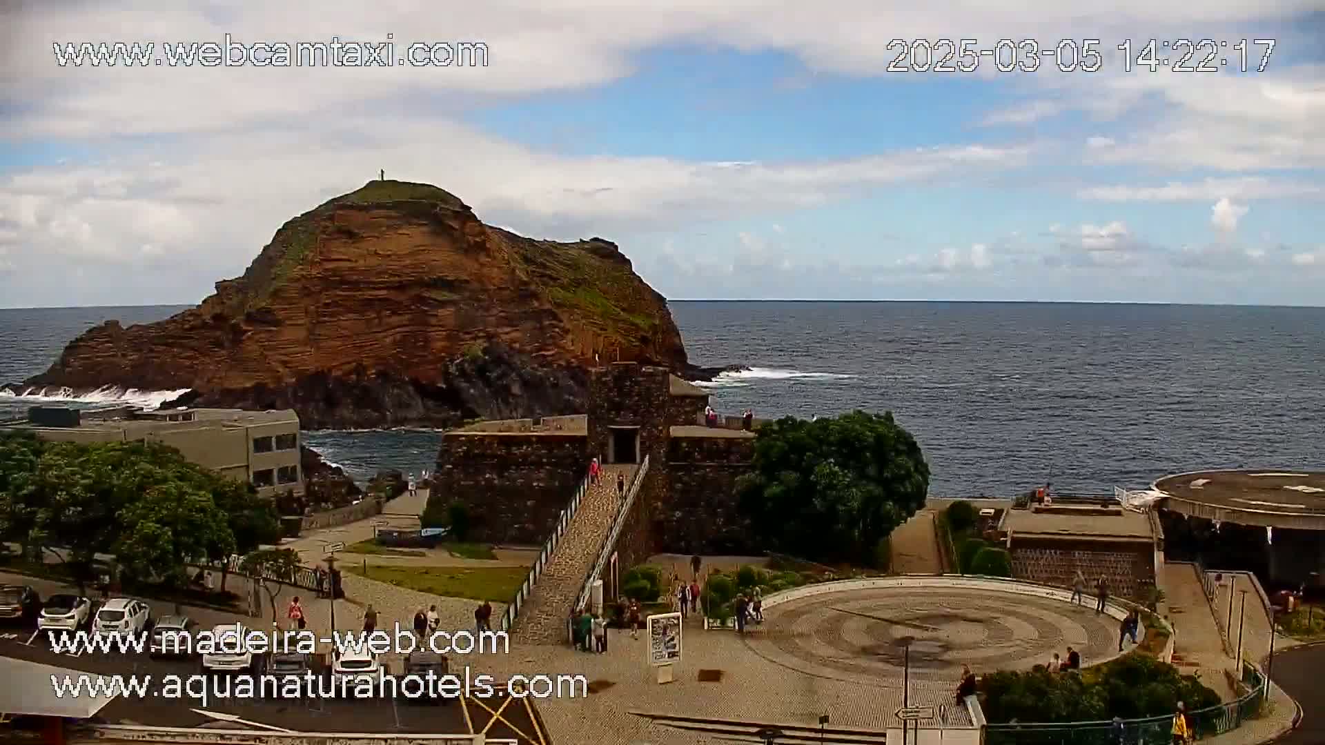 A partly sunny day shows a rocky island and ocean, with a stone structure and a paved area with people near the water.