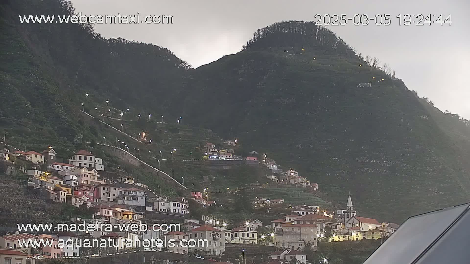 A village of small, brightly lit houses clings to the side of a large, dark green hill under an overcast sky.