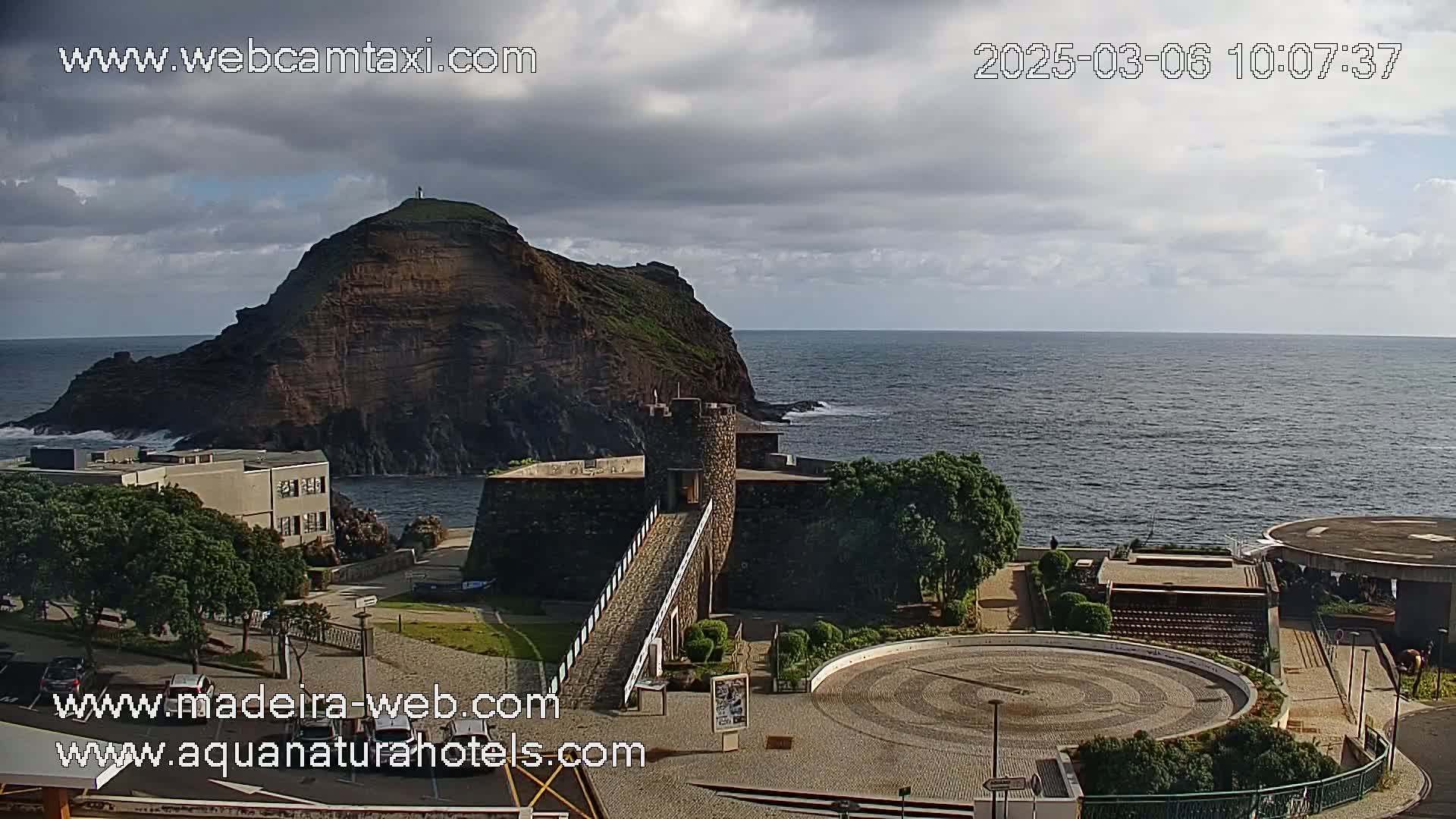 A coastal scene shows a dark, rocky island next to a stone structure and a circular plaza, all under a partly cloudy sky.