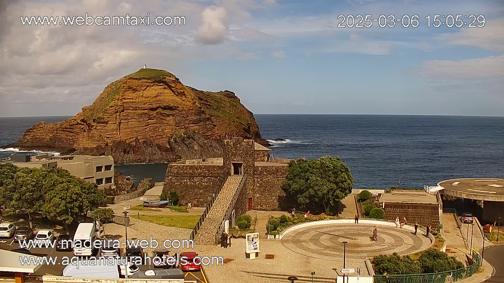 A stone fort with a walkway leading to it sits on a rocky coast next to a steep, reddish-brown cliff overlooking a calm ocean under a partly cloudy sky.