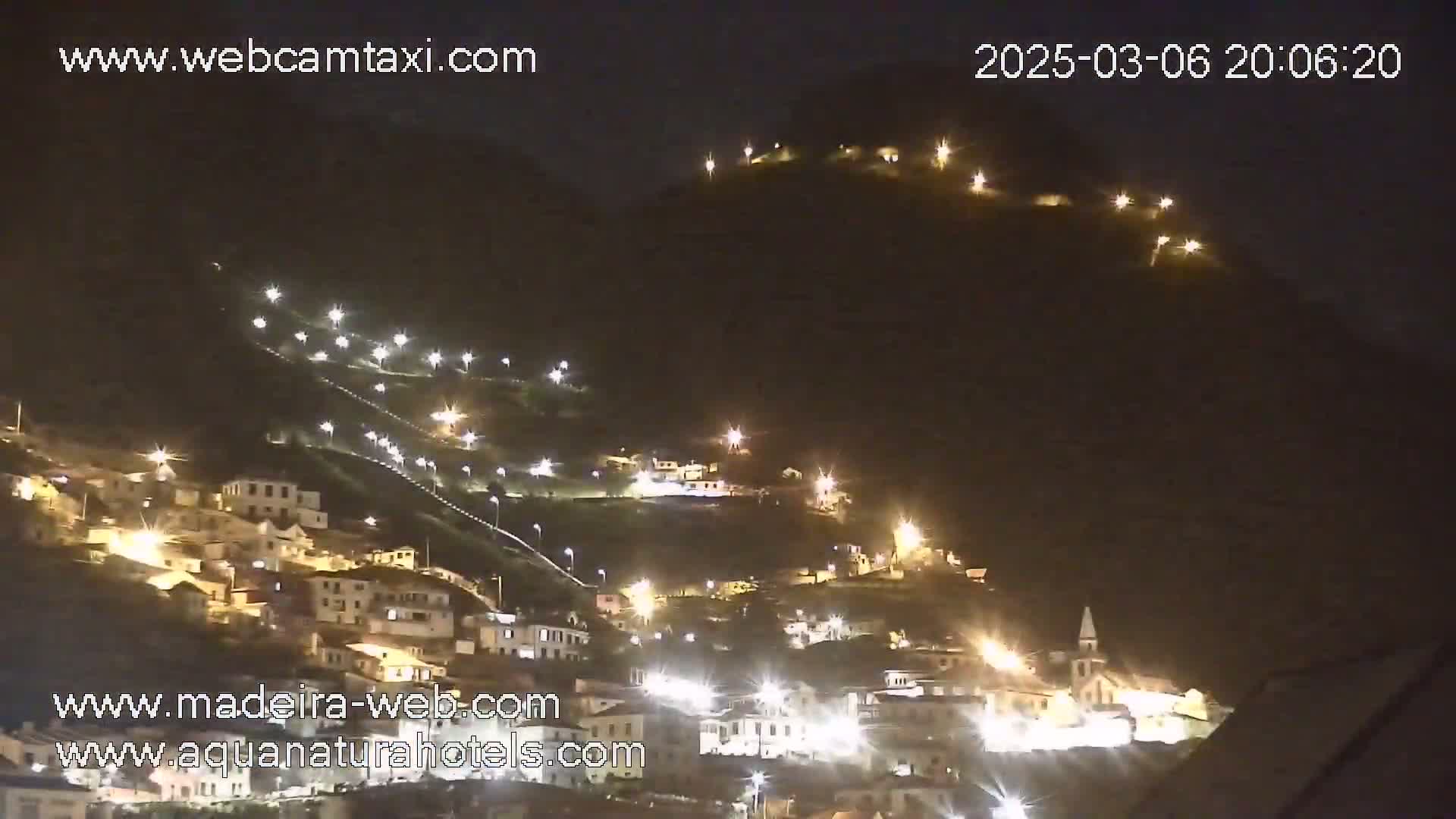 A hillside village is illuminated at night by numerous lights, extending up a steep incline.