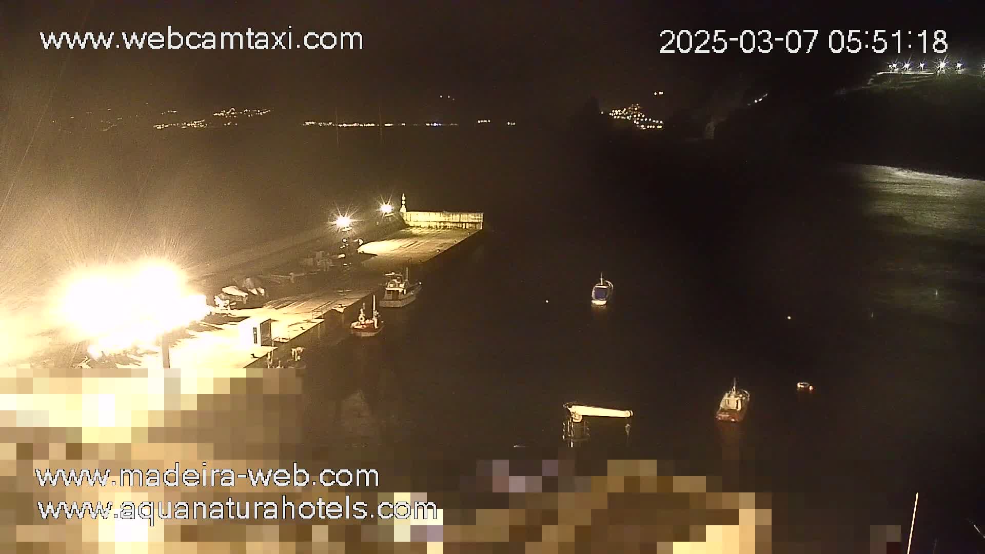 At night, a brightly lit harbor shows several small boats moored at a pier, with a city's lights visible in the distance.