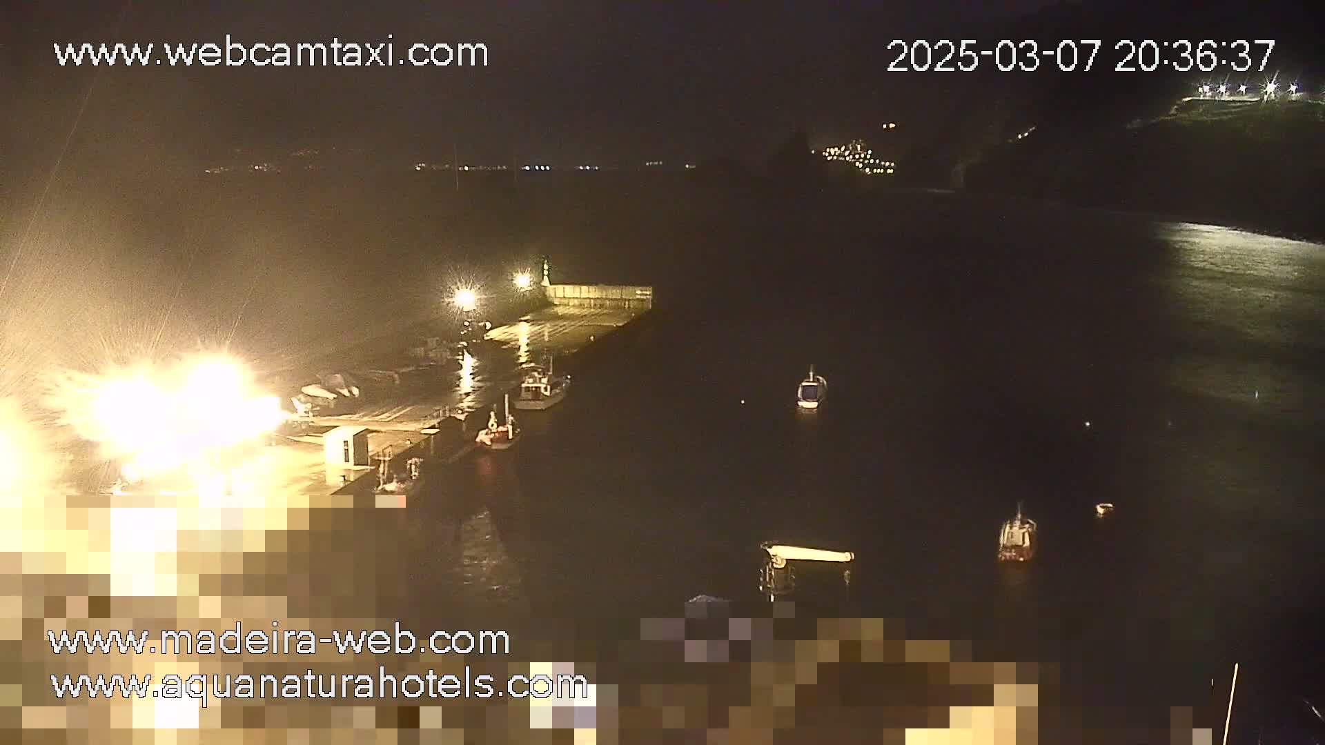 A nighttime harbor scene showing several small boats near a dock, with bright lights illuminating the water and a large light source near the dock.