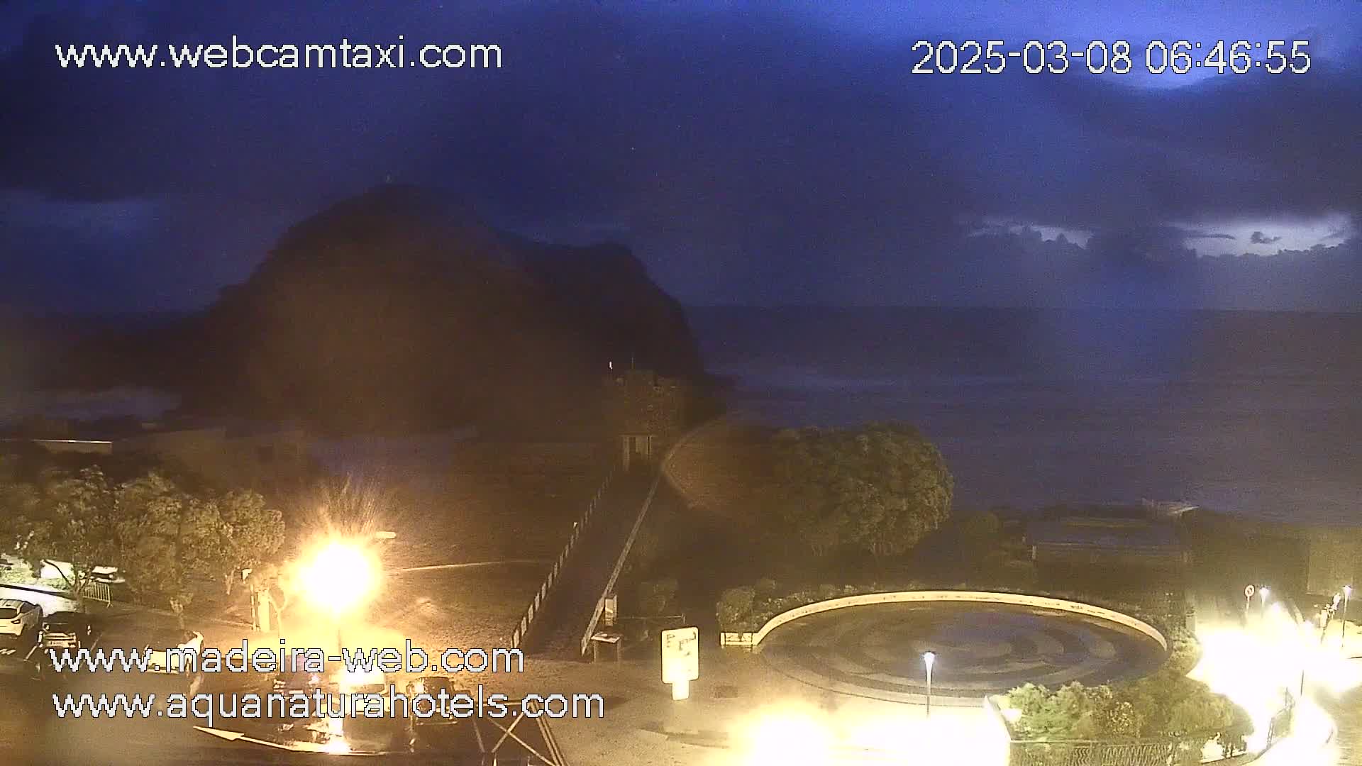 A dark, nighttime coastal scene shows a rocky cliff overlooking a calm sea under a partly cloudy sky.