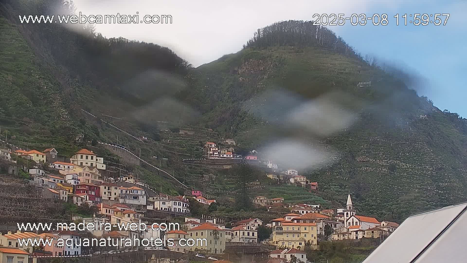 A hillside village with colorful buildings is seen through rain-streaked glass, nestled against a misty green mountain.