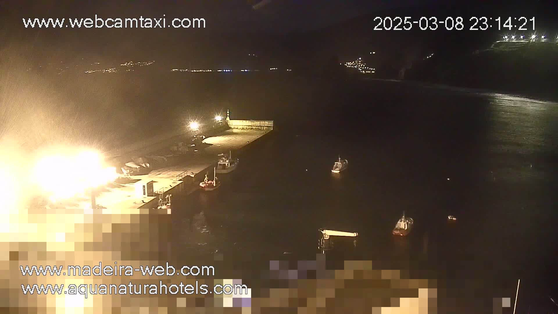 A nighttime view of a harbor with several boats moored alongside a pier, under snowy or hazy conditions.