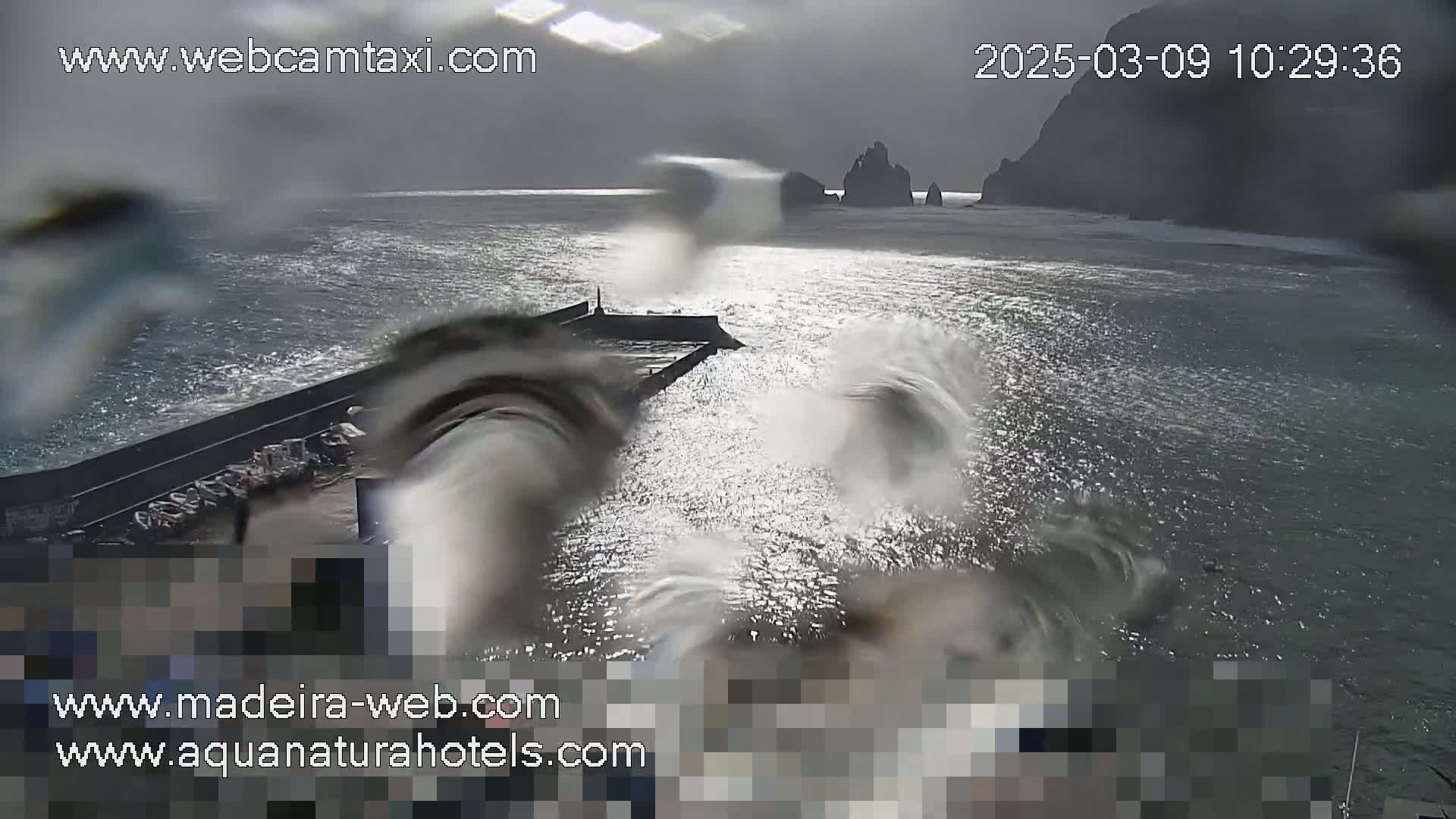 A rain-spattered view shows a choppy sea crashing against a seawall near dark, rocky cliffs under a cloudy sky.