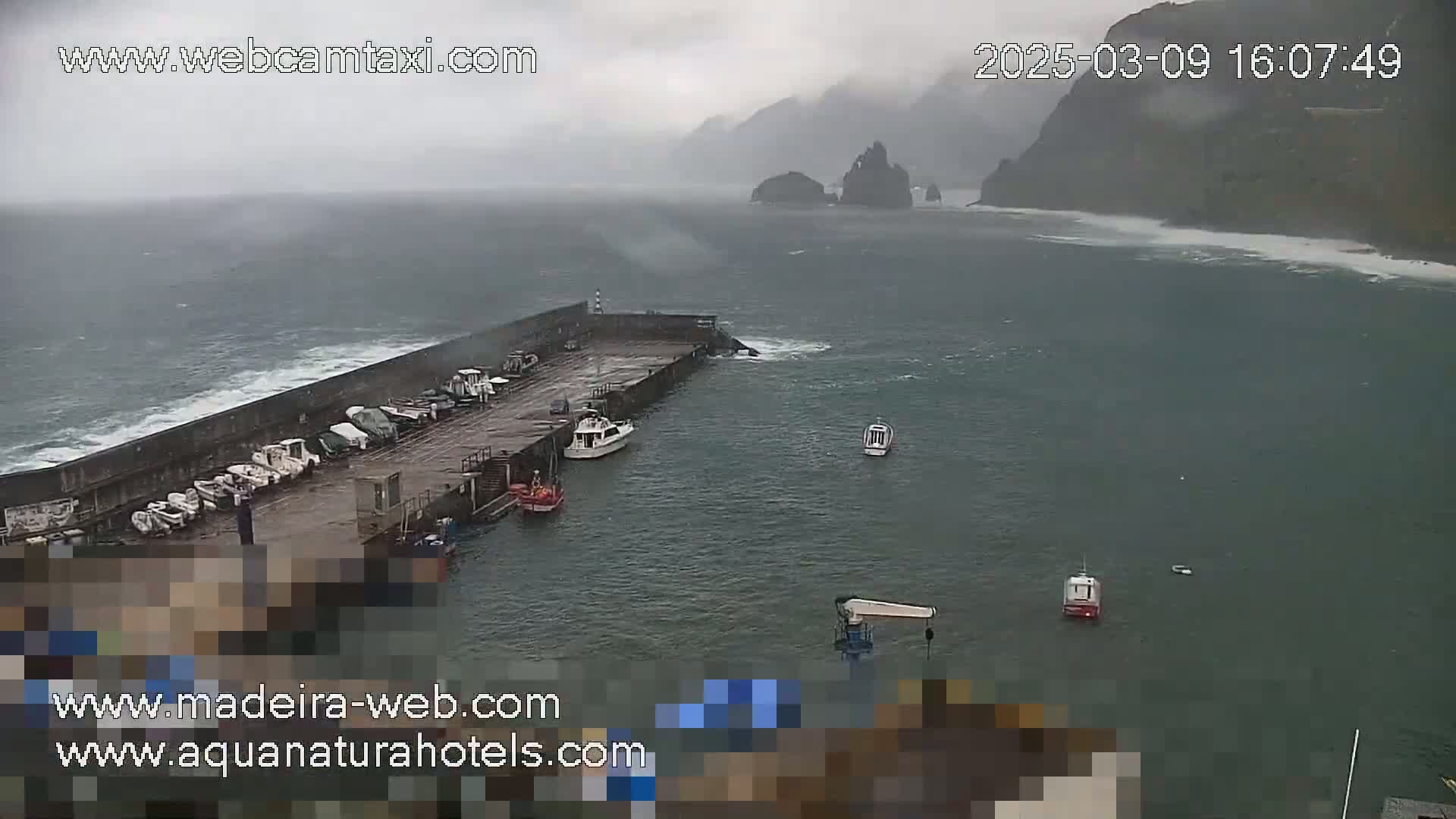 A harbor with several boats docked is seen under stormy, overcast skies, with waves crashing against a seawall near rocky cliffs in the distance.